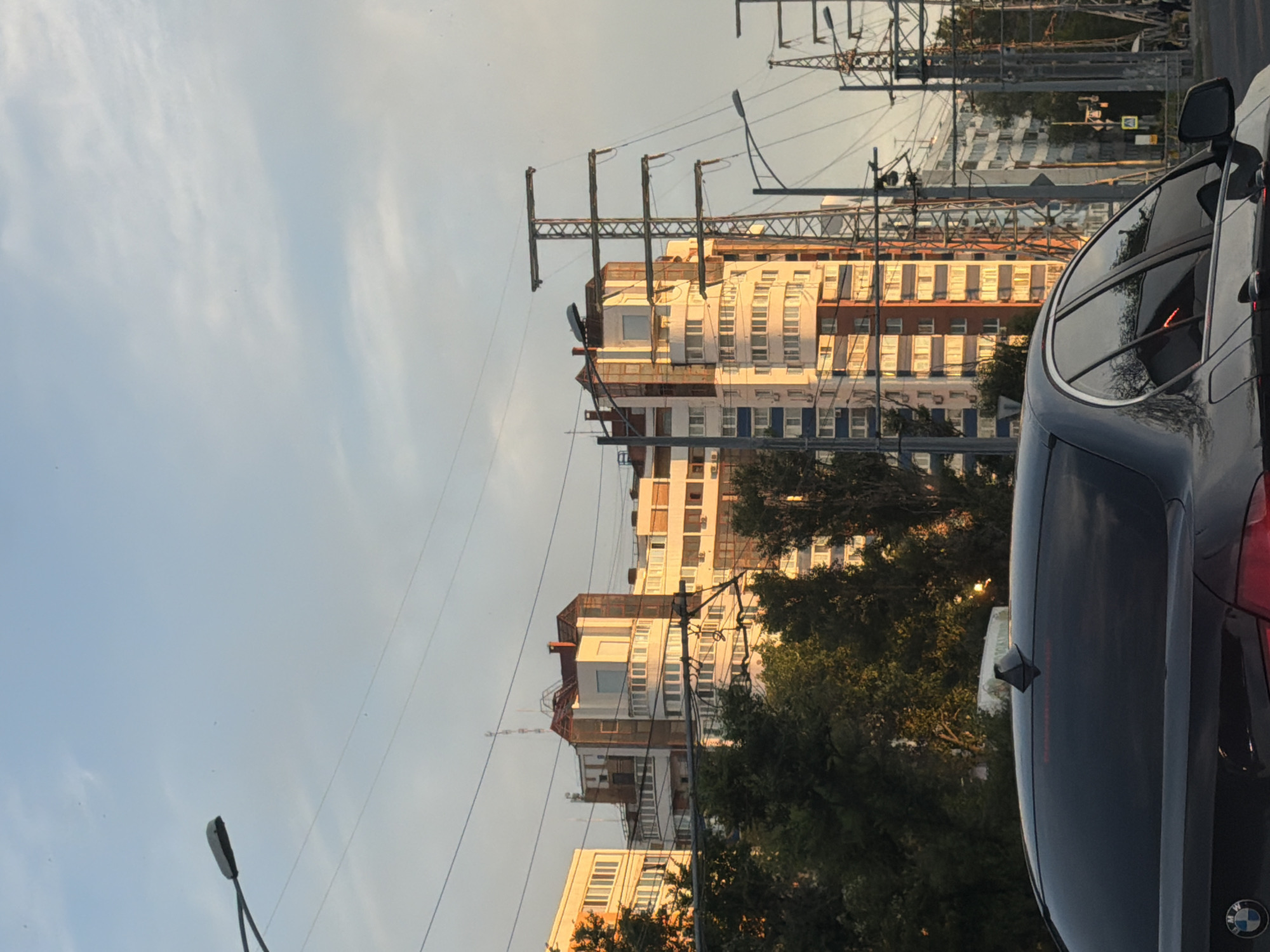 The image shows a city street scene in Samara, Russia, during what appears to be late afternoon or early evening, given the warm, golden light illuminating the buildings. In the foreground, the rear of a dark-colored sedan, possibly a BMW based on the visible badge, is captured. The car has a shark fin antenna on its roof and tinted rear windows. Beyond the car, a line of trees with green foliage is visible.
Dominating the background are several multi-story apartment buildings with a mix of architectural styles. Some buildings have prominent balconies and numerous windows. The structures are bathed in the golden sunlight, creating highlights and shadows. Interspersed among the buildings and trees are tall power line towers and numerous overhead wires, indicating an urban electrical infrastructure. Streetlights are also visible along the road. The sky above is a pale blue with some scattered clouds. The overall impression is a typical view of residential architecture and infrastructure in a Russian city.