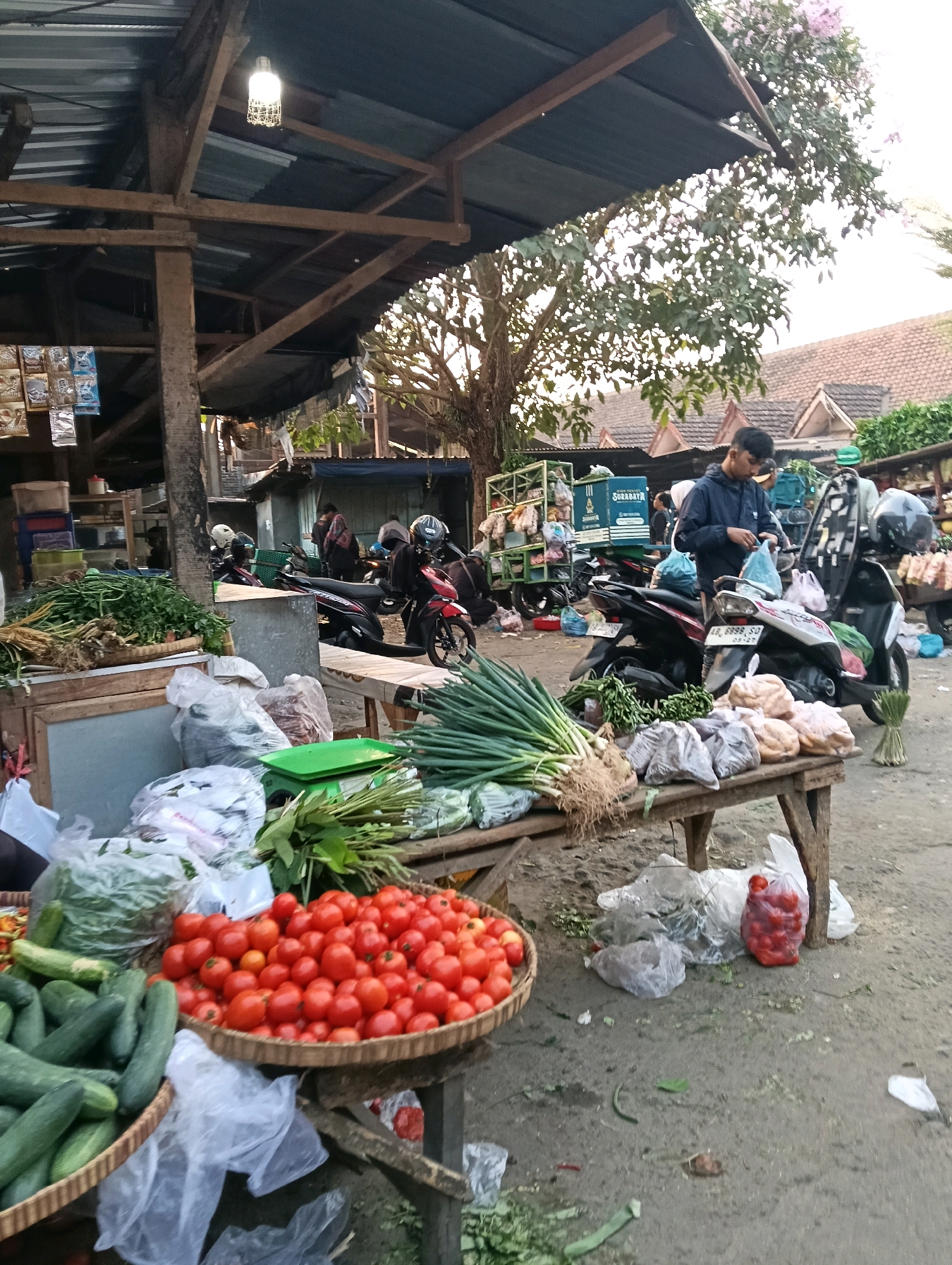 The image depicts an outdoor market scene in Magelang, Indonesia. In the foreground, a wooden table displays an assortment of fresh produce. A large woven basket is filled with ripe red tomatoes. To the left of the tomatoes, another woven basket contains numerous cucumbers. Bunches of green onions and other leafy vegetables are also visible on the table. Several plastic bags, some containing produce and others empty, are scattered around the display. In the background, a person wearing a dark jacket and a black cap is standing near a motorcycle, appearing to be engaged in a transaction or preparing to depart. Other motorcycles are parked in the vicinity, some with helmets on their seats. The market stalls are constructed from wood and corrugated metal, with tarps providing shade. Behind the stalls, traditional Indonesian houses with tiled roofs are visible, along with trees and foliage. The ground is dirt or gravel, with some debris present. The overall impression is of a bustling local market in Indonesia.
