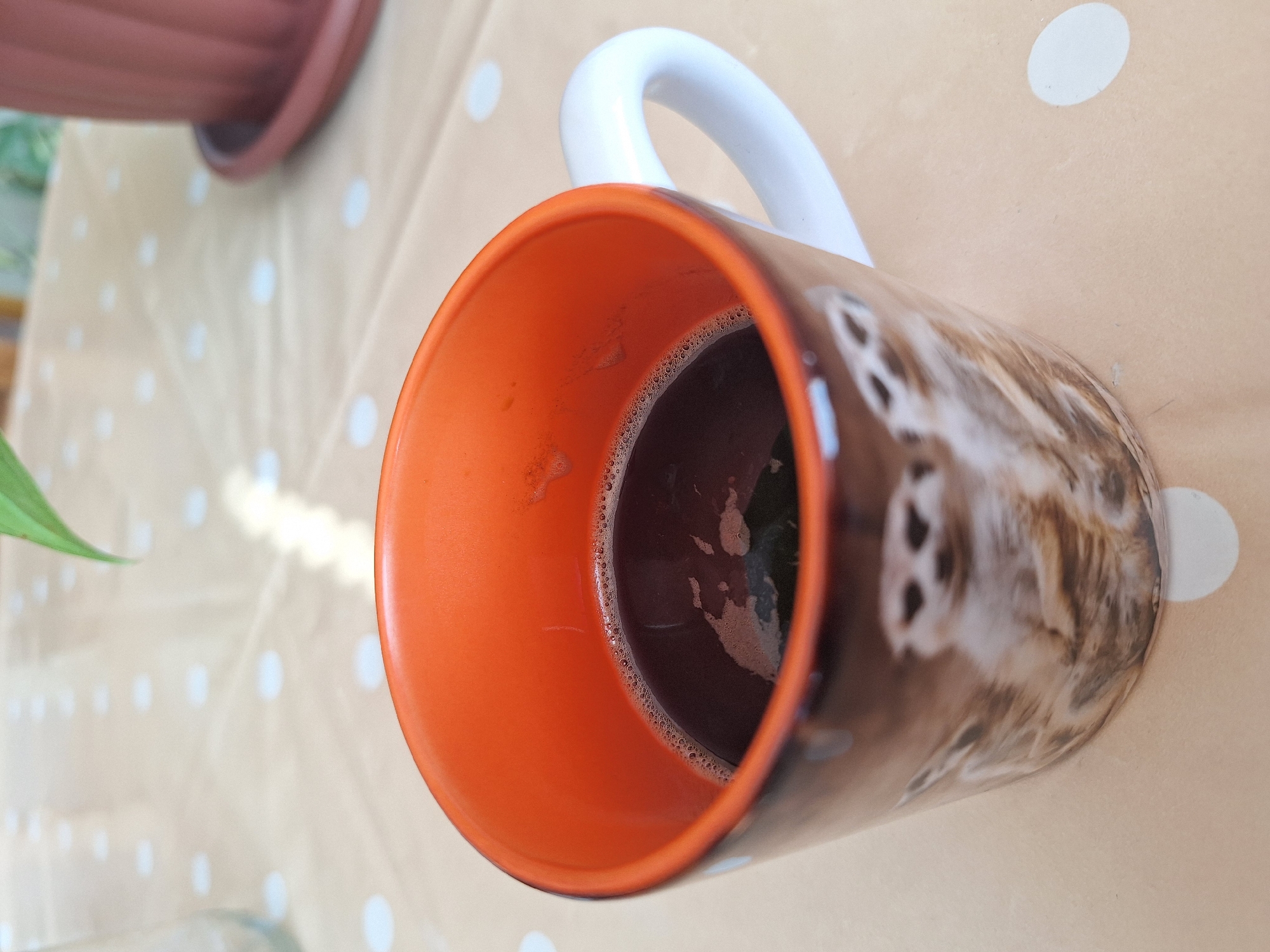 The primary subject of this image is a coffee mug, filled with a dark beverage, likely coffee, topped with a layer of small bubbles. The mug's interior is a bright orange, contrasting with its white handle. The exterior features a brown and white furry animal print, possibly depicting a cat or dog, with distinct patterns.
The mug is placed on a light beige or cream-colored surface adorned with a pattern of white polka dots, which appears to be a tablecloth or mat. In the upper left background, part of a reddish-brown, fluted plant pot is visible. A vibrant green plant leaf peeks into the lower left corner of the frame.
The lighting suggests the photo was taken during the daytime, with natural light creating subtle highlights and shadows. There are no people visible, and no specific event or activity is taking place beyond the presence of the drink. The scene conveys a calm, domestic setting, potentially a home in Montana, Bulgaria. There is no visible text in the image.