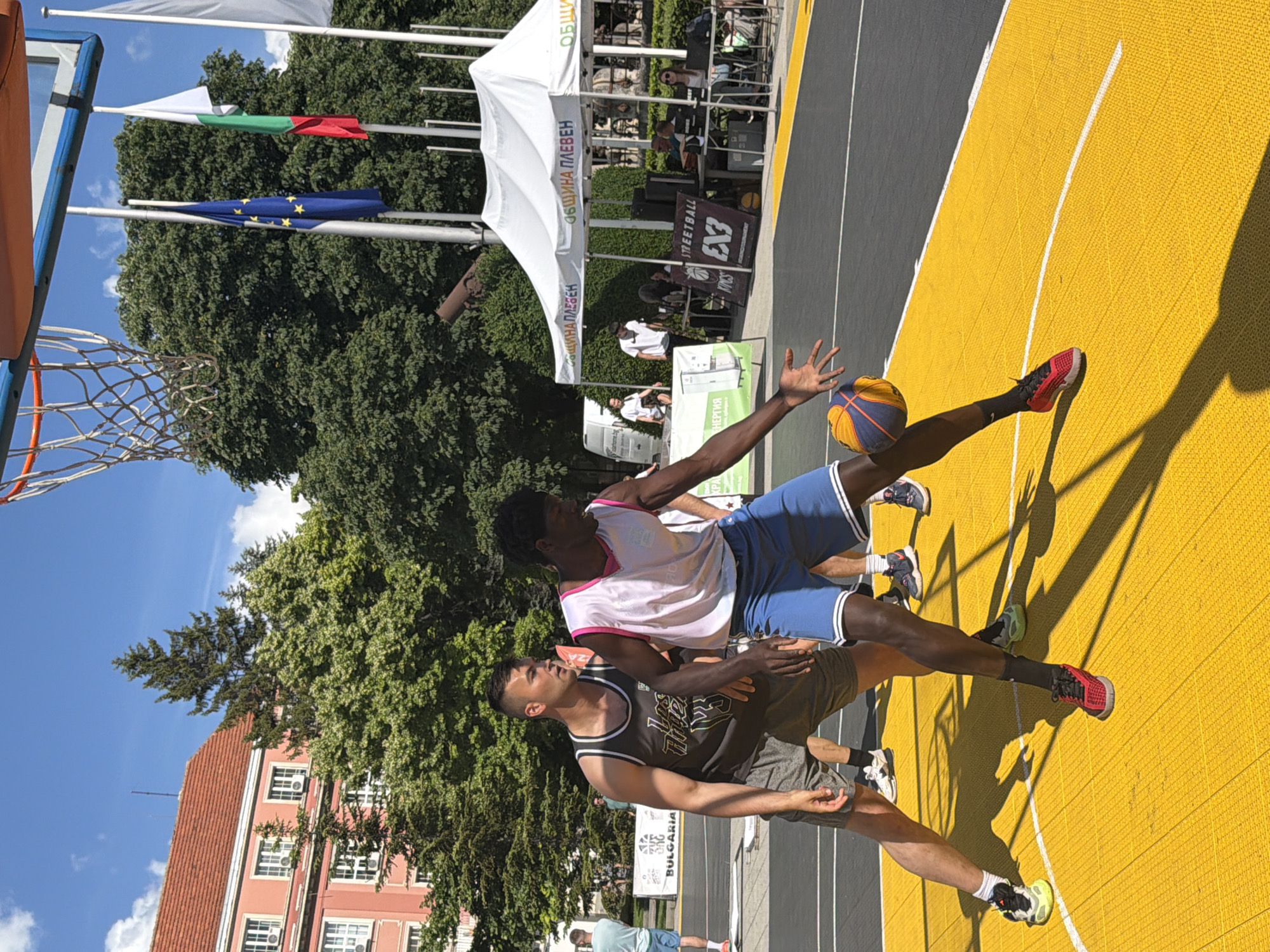 Two male basketball players, one Caucasian and one African American, are engaged in a game on a bright yellow outdoor court. The Caucasian player, wearing a dark grey tank top with "Los Angeles" text and grey shorts, is positioned to the left, looking intently at the ball. The African American player, in a white tank top with pink trim and blue shorts, is on the right, dribbling a basketball with his left hand, his body angled towards the basket. Both players are wearing athletic shoes. In the background, a building with a red-tiled roof and white-framed windows is visible, along with lush green trees. Several white tents are set up, one with Bulgarian text reading "ОБЩИНА ПЛЕВЕН" (Municipality of Pleven). Flags, including the European Union flag and the Bulgarian flag, are flying on poles behind the tents. The scene suggests a public outdoor event or competition in Pleven, Bulgaria.