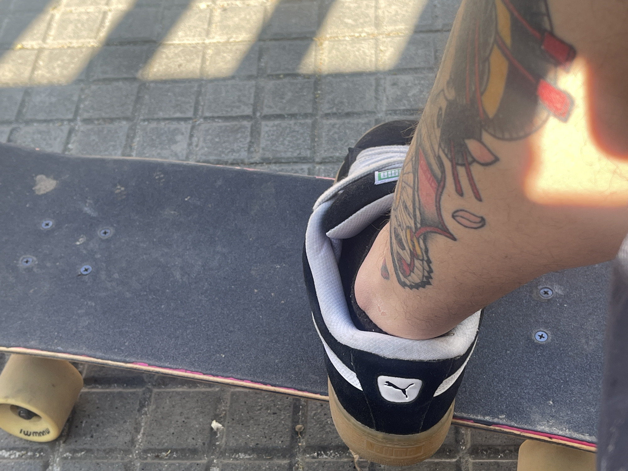 A person wearing a black and white Puma suede shoe with a gum sole stands on a skateboard on a sidewalk. The person has a tattoo on their ankle. The skateboard is black and has yellow wheels. The sidewalk is made of gray square tiles. The location is Cerdanyola Del Vallès, Spain.