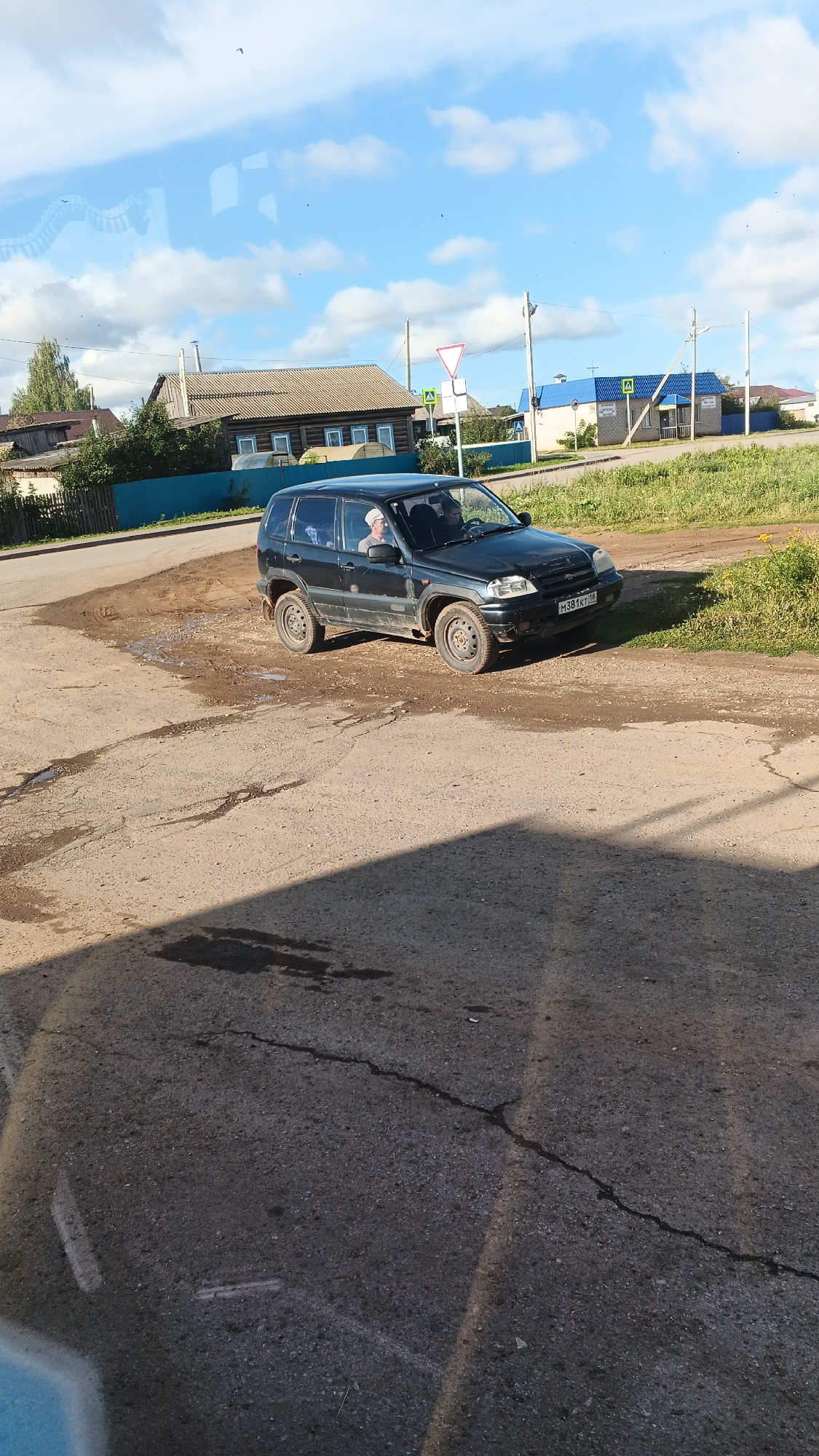 A dark-colored SUV is parked on a dirt and asphalt road in a rural setting. The vehicle is facing slightly to the right of the camera. A man wearing a light-colored cap and a light-colored shirt is visible in the driver's seat, looking towards the right. Another person is partially visible in the passenger seat. The road surface is uneven with patches of mud and puddles. In the background, there are several wooden houses with tiled roofs, a blue fence, and green grassy areas. The sky is blue with scattered white clouds. There are traffic signs visible, including a triangular yield sign and a square sign with a yellow triangle and a black exclamation mark. The license plate of the vehicle is visible as "M 381 KT 18". The scene appears to be in the village of Kiyasovo, Russia.