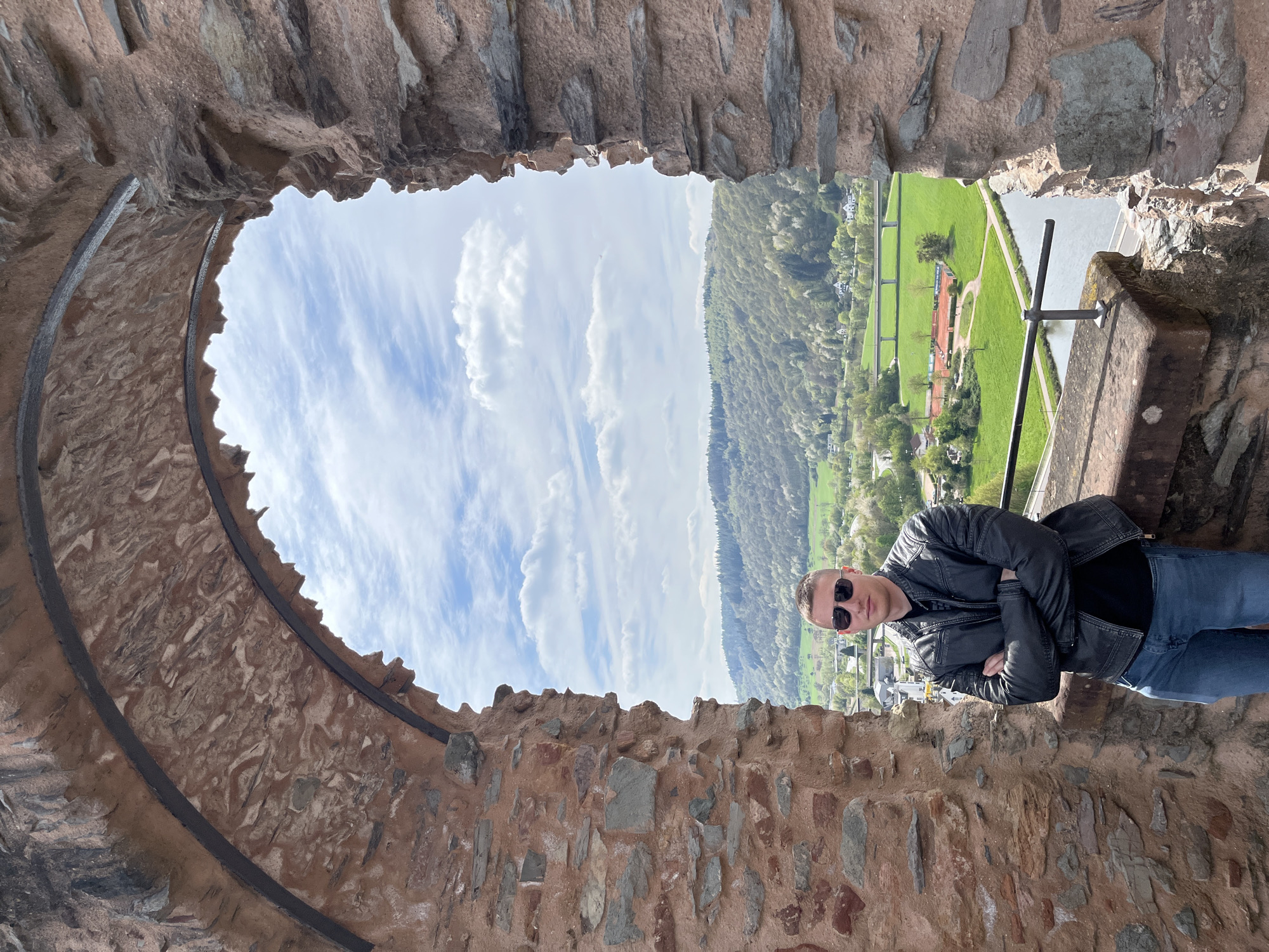 A man stands in an arched window of an old stone structure, likely a castle or historical building. He is wearing a black leather jacket, sunglasses, a black shirt, and blue jeans. He has his arms crossed. Outside the window, a landscape is visible, showing green fields, a river, trees, and buildings. The sky is partly cloudy. The structure is identified as being in Saarburg, Germany, based on visual characteristics of the landscape and architecture.