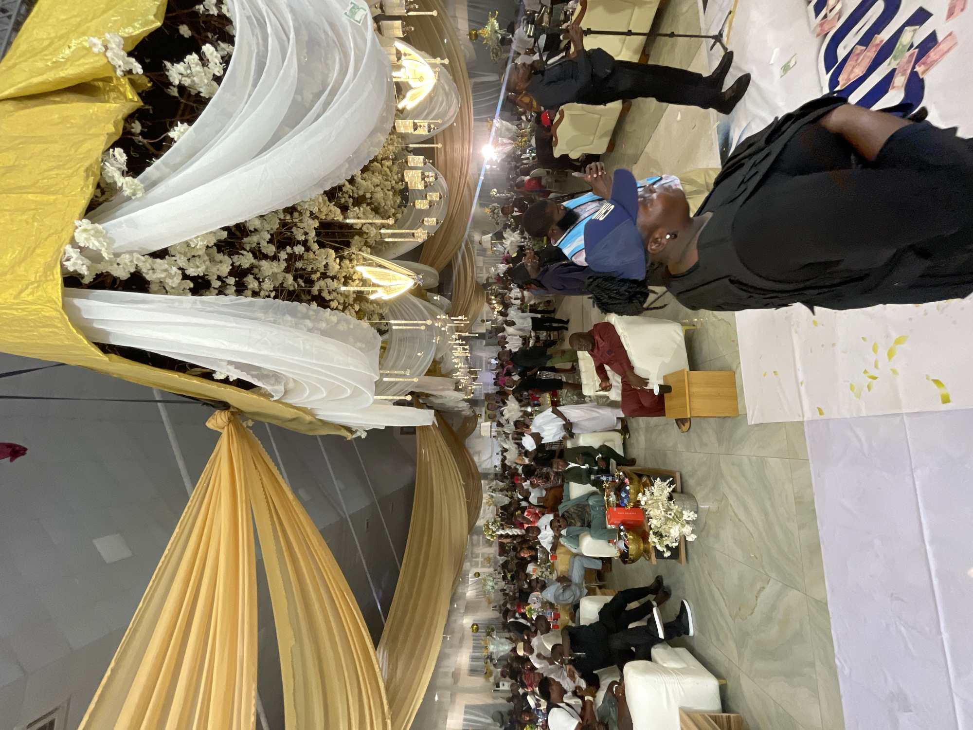A gathering of people is taking place in what appears to be a wedding reception hall in Barikin Jirga Summa, Nigeria. The hall is decorated with gold and white drapery, floral arrangements, and decorative lighting. Many attendees are seated at tables and appear to be observing a ceremony or event. Several people are standing around with various expressions. The scene appears active and social.