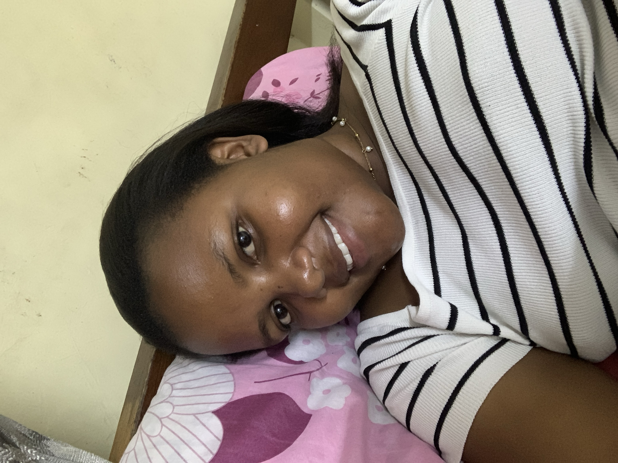A dark-skinned woman is lying on a bed. The woman is resting her head on a pink pillow with white floral patterns and maroon heart-shaped designs. She has dark, straight hair pulled back from her face, and she is wearing a gold necklace. The woman is smiling and looking towards the camera. She is wearing a white and black striped shirt. A wooden headboard is visible behind her. A cream-colored wall is in the background. It is not possible to determine the location of this photo as Dar es-Salaam, Tanzania with the given information.
