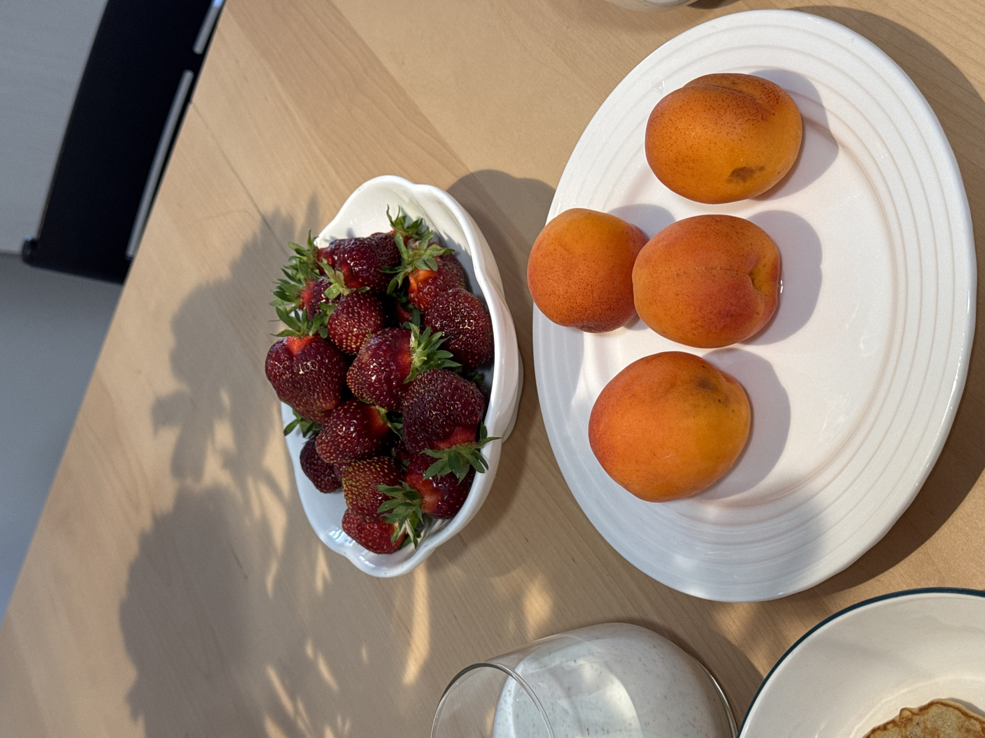 A close-up, overhead shot captures a wooden table with a spread of fresh fruit and a drink. In the center, a white bowl overflows with ripe strawberries. To the right, a white plate holds three apricots. A clear glass to the left of the strawberries contains a milky white beverage. The background shows the shadow of a chair on the table. The scene is set in an indoor location, likely a home or restaurant, and there is no discernible indication of its location within Izhevsk, Russia.