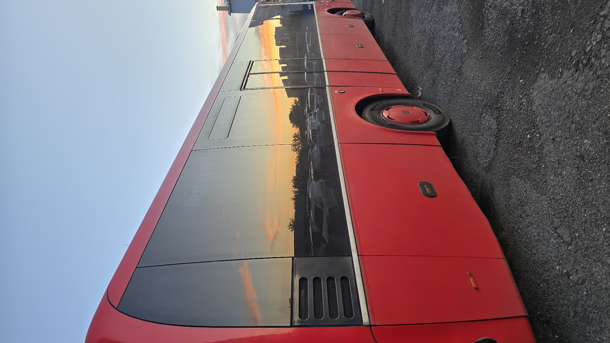 A side profile of a red bus is shown parked on an asphalt surface. The bus has large tinted windows that reflect the sky, trees, and buildings, suggesting it is sunset or sunrise. The sky is a clear blue with hints of orange and pink clouds near the horizon. In the reflection, several cars are visible, appearing to be in a parking lot. The bus itself is clean and appears to be in good condition. There are no people visible in the image. The environment appears to be an outdoor lot, possibly a parking area or depot, with the presence of buildings in the background reflection. The image does not contain any specific landmarks that would indicate its precise location in the city of Kyiv, Ukraine.