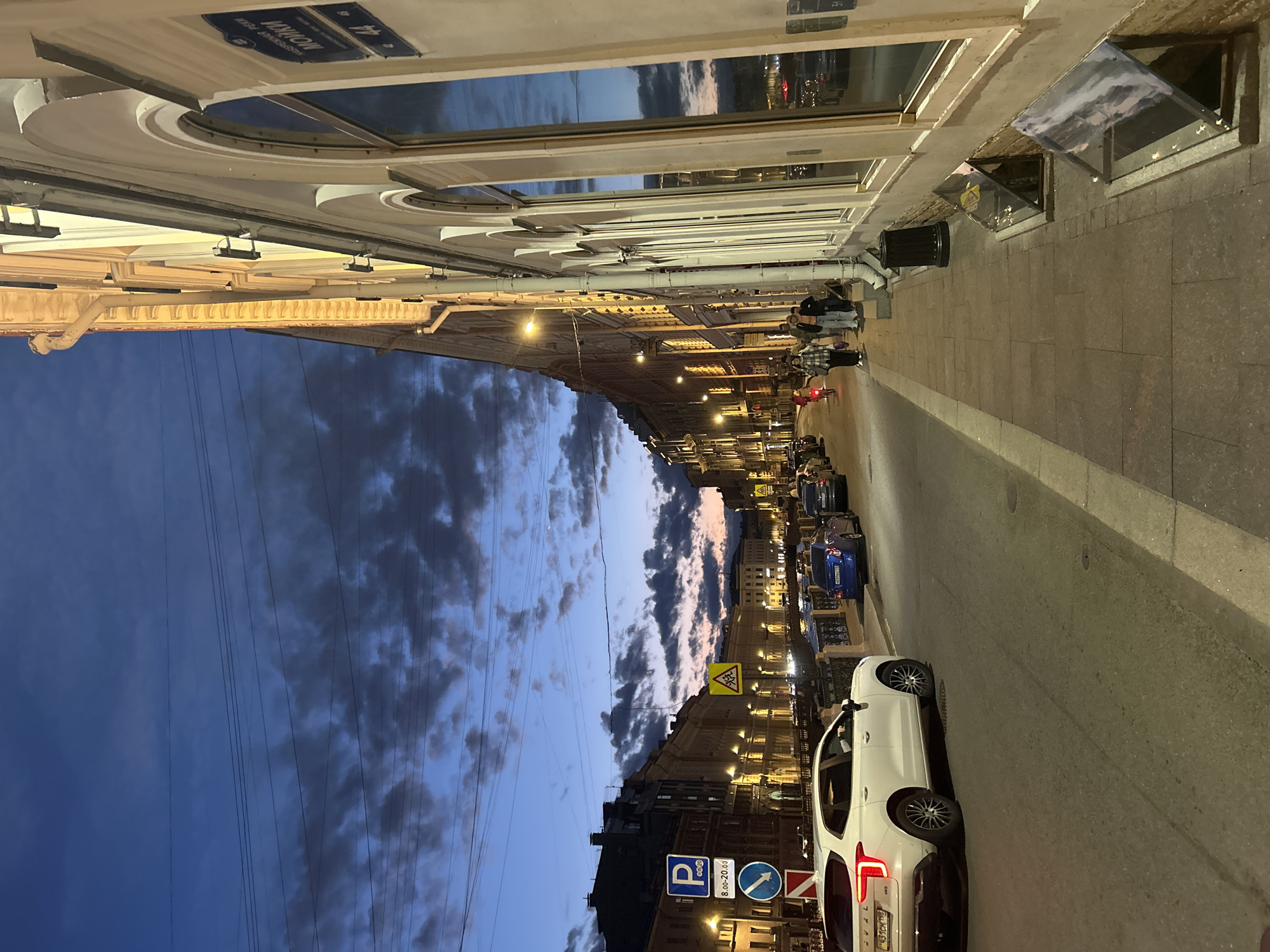 The image depicts a street scene at dusk in a city. The sky is a deep blue with scattered dark clouds, transitioning to lighter hues near the horizon, suggesting the last moments of daylight. A white SUV is parked on the left side of the street, facing away from the camera, with its rear visible. Further down the street, other cars are parked and driving, illuminated by streetlights and building lights.
On the right side of the image, a light-colored building with arched windows and ornate architectural details runs along the sidewalk. The building is lit, and its windows reflect the dusky sky. A street sign is attached to the building, with Cyrillic text visible. Pedestrians are walking on the sidewalk in front of the building.
The street itself is paved, and there is a sidewalk made of stone slabs. The environment appears to be an urban setting with a canal or river running parallel to the street, bordered by a decorative railing. The overall atmosphere is calm and tranquil, with the city lights beginning to glow as night approaches.