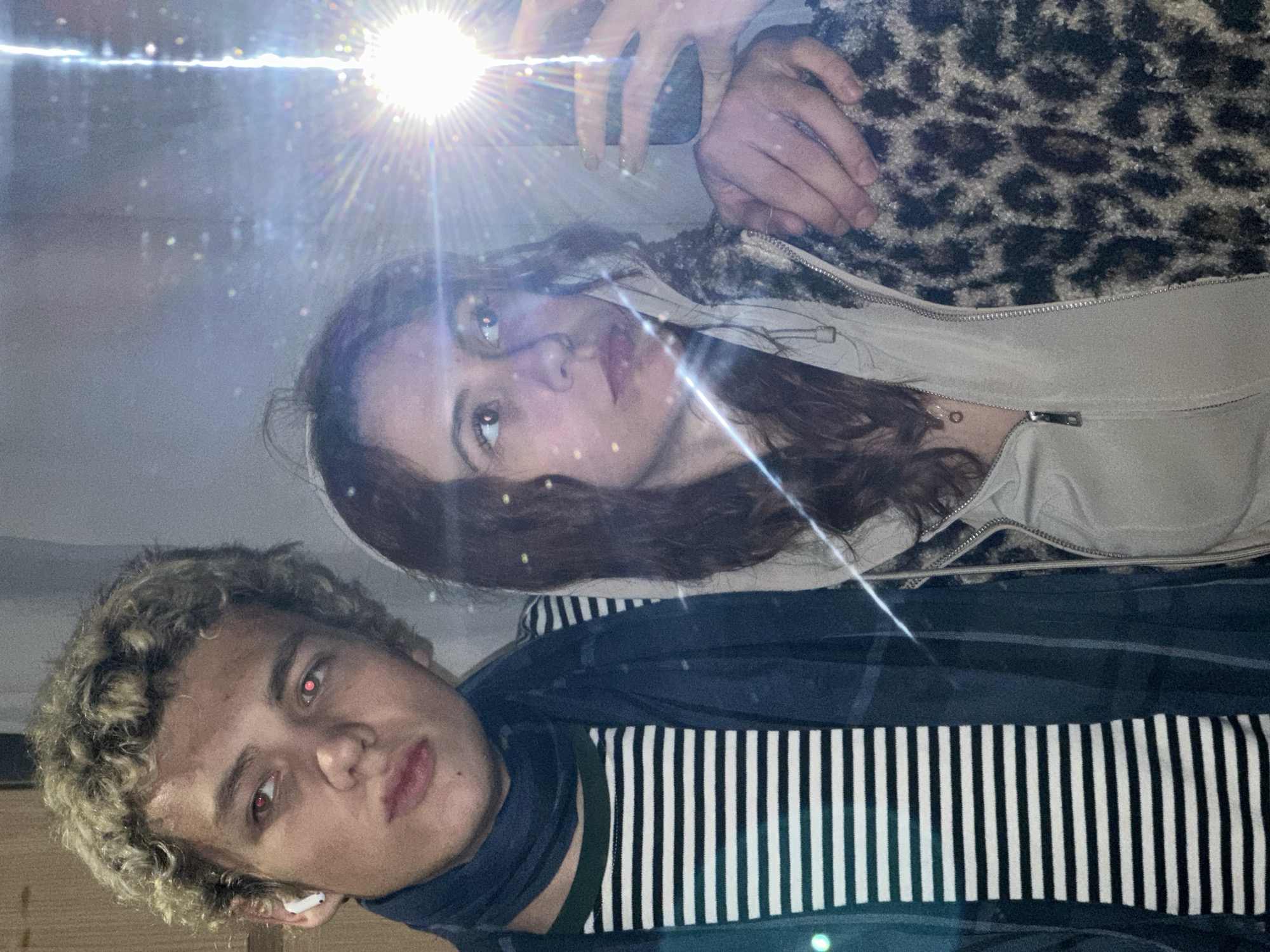 This image captures a close-up selfie of two young individuals, a female and a male, taken indoors in front of a reflective surface such as a mirror or window, likely at night given the use of a bright camera flash. The photograph is presented rotated 90 degrees clockwise from its original orientation.
The female is prominently featured in the upper portion (when viewed upright). She has long, dark, wavy hair and wears a light-colored, possibly beige or off-white, top beneath a textured, brown and black leopard print jacket. Her hands are visible holding a smartphone, from which a powerful flash emanates, creating a strong starburst effect in the upper left of the image and illuminating the scene. She looks slightly upwards with a neutral to contemplative expression.
The male is positioned below the female, to her right (when viewed upright). He has short, curly blonde or light brown hair and is looking directly at the camera. His eyes show a distinct red-eye effect from the flash. He wears a dark blue or black top, possibly a turtleneck or scarf, over a black and white vertically striped shirt. A white earbud is visible in his right ear.
The background is dark and indistinct, with scattered bright specks that could be dust on the reflective surface or light reflections. The atmosphere is casual and informal. The location context provided indicates this moment occurred in Novi Sad, Serbia, although no specific visual cues within the image confirm this. There is no visible text in the image.