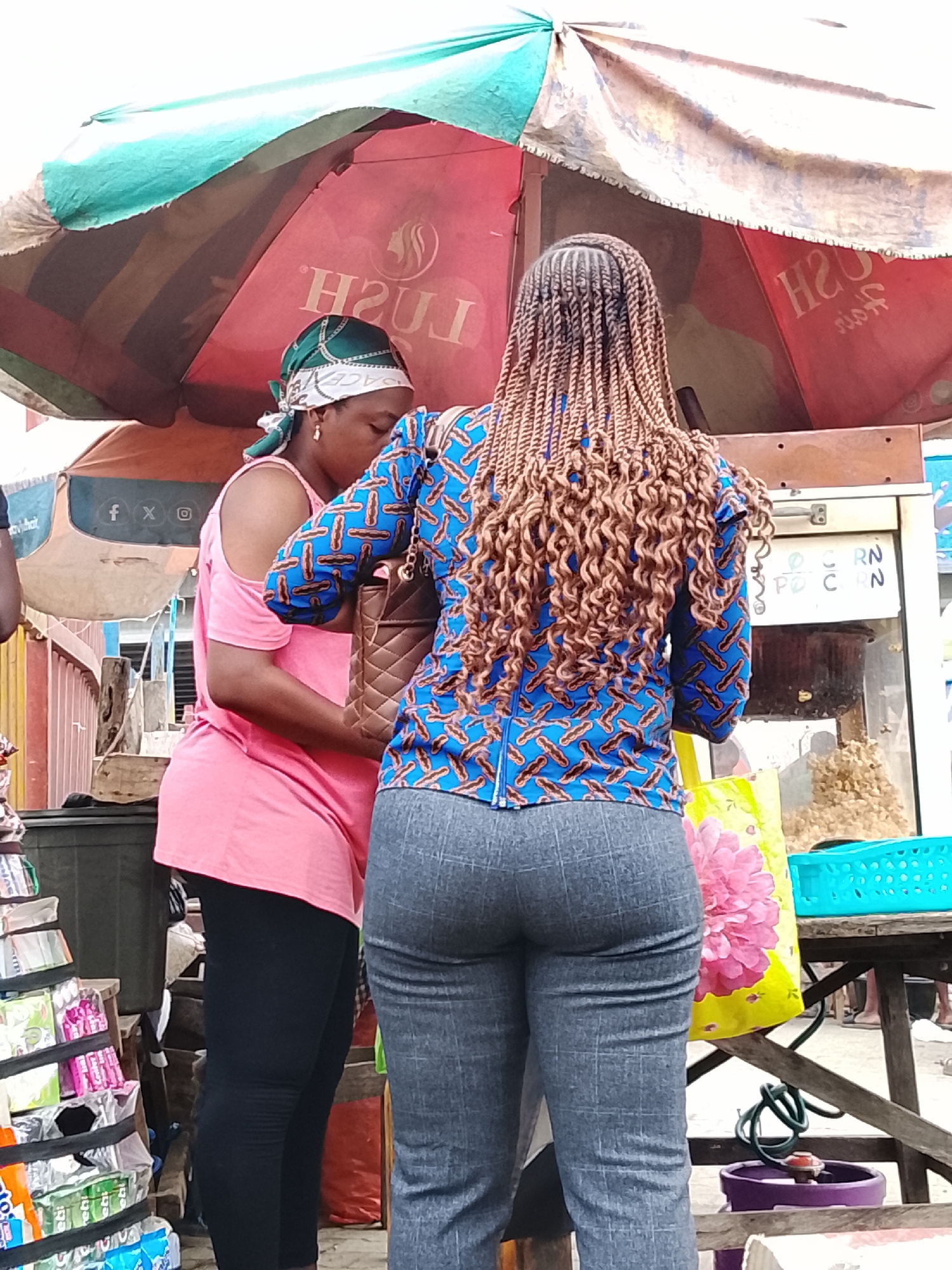 The image shows two women standing outdoors at a market stall. The woman on the left, wearing a pink sleeveless top and black leggings, has a green and white headscarf. She is holding a quilted brown handbag. The woman on the right has her back to the camera, wearing a blue patterned jacket with a zip-front and grey checkered trousers. Her hair is styled in long, intricate braids. Behind them, a popcorn machine is visible, with popcorn in the glass container and more popcorn on display on a wooden table. A large, colorful umbrella is overhead, providing shade. Various items are displayed on shelves to the left of the frame. The scene appears to be in a bustling urban environment, likely a street market in Lekki, Nigeria.