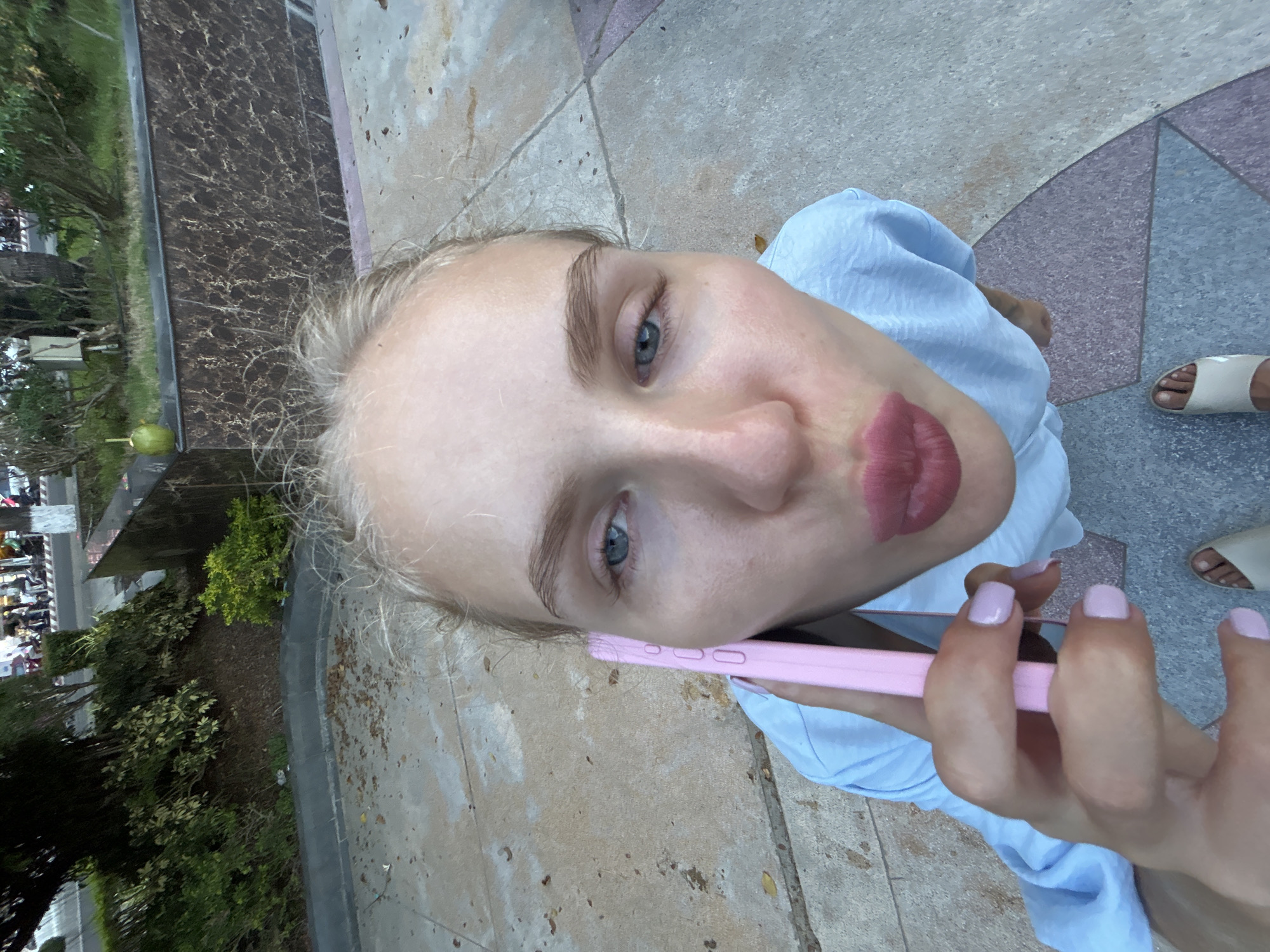 A close-up, overhead shot captures a blonde woman with a light blue shirt holding a pink smartphone to her ear, her lips pursed. She is standing on a paved area with patterned sections, possibly in a public square or walkway, near a street with passing vehicles and pedestrians. Lush green foliage and a dark, marbled wall form the background. The scene suggests an urban outdoor setting in Nha Trang Ward, Vietnam.