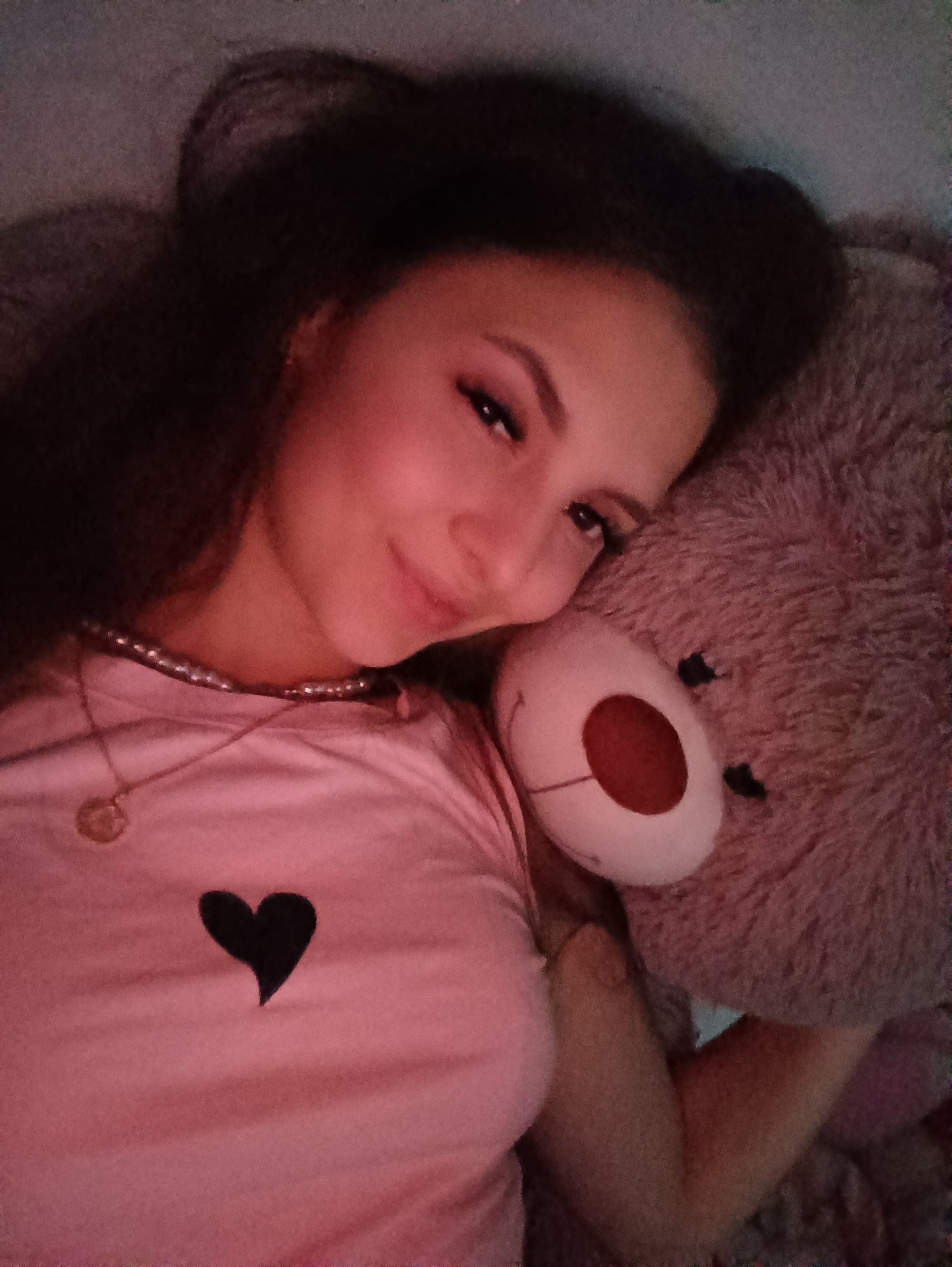 A woman is lying down next to a stuffed teddy bear. She is wearing a pink t-shirt with a black heart on it, a pearl necklace, and a gold pendant. Her dark hair is styled in buns on the top of her head. She is looking at the camera with a slight smile. The teddy bear is light brown and has a red nose. The background is blurry. The location is unknown.