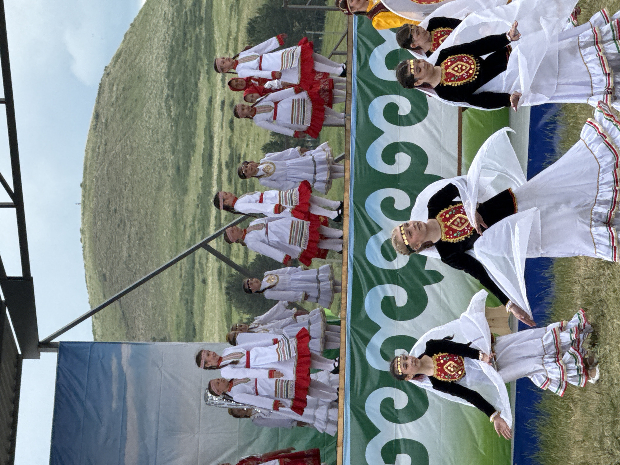 The image depicts a cultural performance taking place outdoors in Nurkeevo, Russia. A group of young girls, dressed in traditional folk costumes, are performing on a stage. The costumes consist of white dresses with red and green trim, and some girls wear headbands. The backdrop of the stage features a blue sky with clouds, and behind the stage, a green, grassy hill is visible under a cloudy sky. In the foreground, another group of girls, also in traditional attire, are dancing. Their outfits include black tops with ornate gold and red embroidery, and long, flowing white skirts with ruffled hems. They are also wearing head adornments. The ground is covered in grass, and the overall scene suggests a festive cultural event.