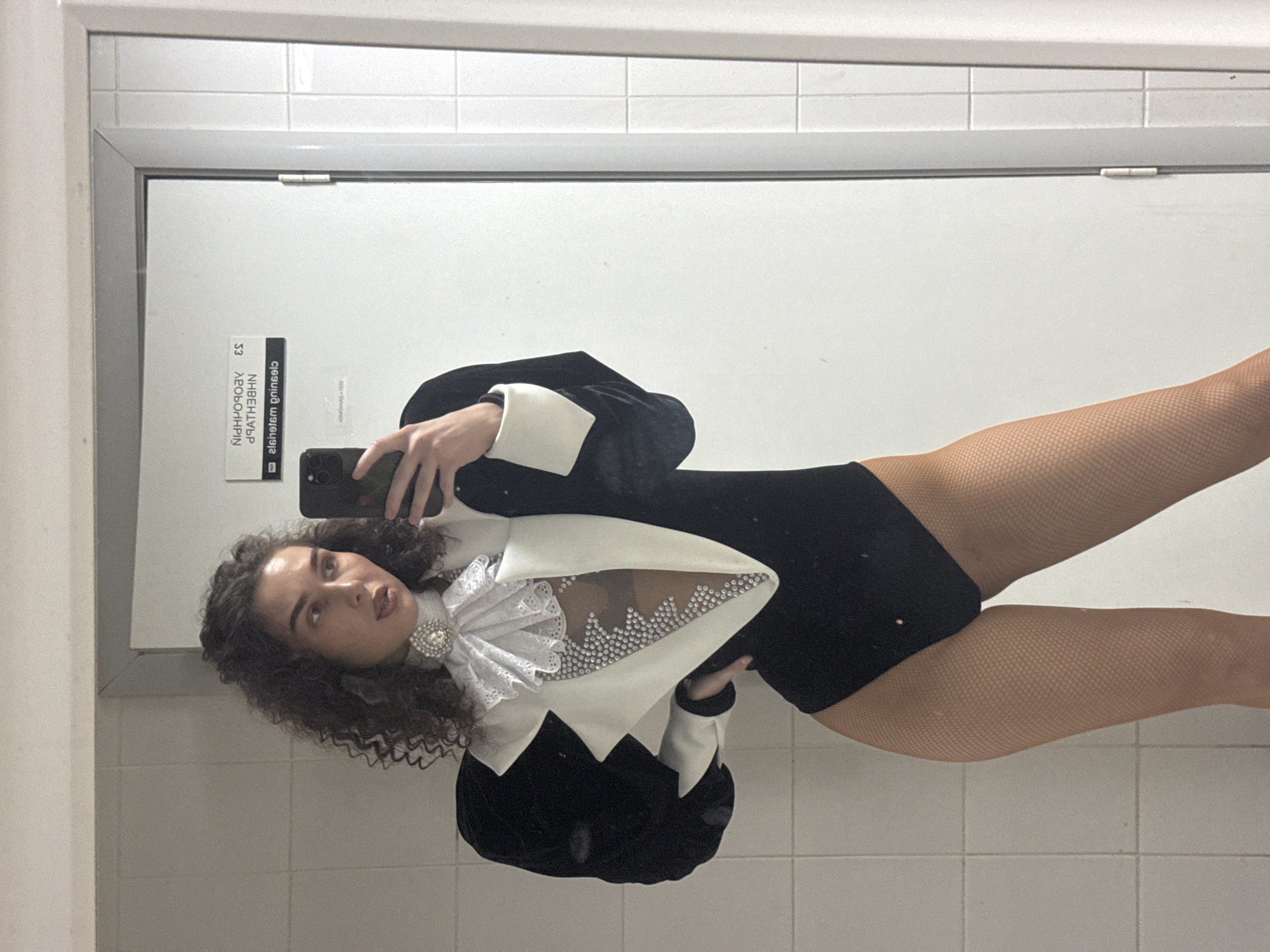 A person stands in front of a tiled wall and a closed door, taking a mirror selfie. The person is wearing a black leotard with a dramatic white collar and lapels adorned with rhinestones. The leotard has puffed black velvet sleeves with white cuffs. They are also wearing fishnet tights. The background appears to be a restroom or dressing room. There is a sign on the door with text in Cyrillic, which reads "ЕСИЗ" and "NHB3HL38". There is also text in Latin script on a sticker on the door that reads "cleisielsm qninsels". The person is holding a smartphone in their right hand to take the photo.
