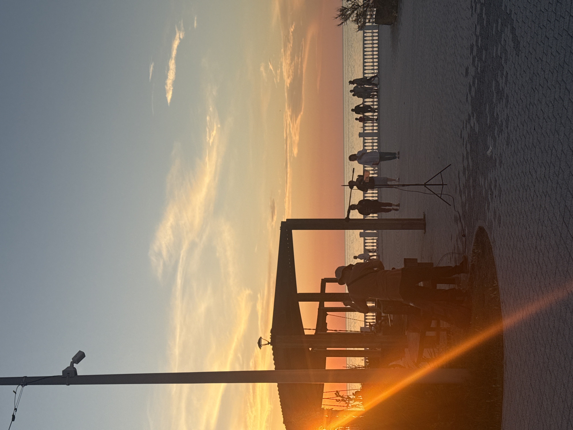 The image depicts a sunset scene on a paved promenade next to the sea. A person wearing a hat is standing in the foreground, facing away from the camera, appearing to be setting up equipment that includes a tripod. Behind them, a covered structure with wooden supports is visible. Several other individuals are walking and standing along the promenade, some silhouetted against the vibrant orange and yellow sky. The sea is calm, and a white balustrade runs along the edge of the promenade. A lamppost with a security camera is on the left side of the frame. The sun's rays create a strong lens flare that cuts diagonally across the left portion of the image.