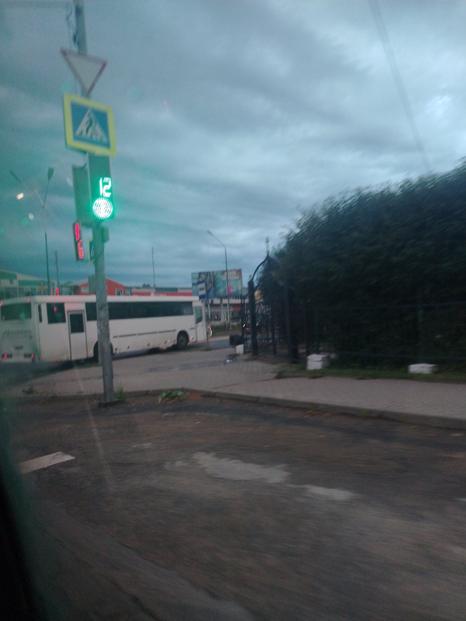 The image shows a street scene in an urban environment under a cloudy sky. A white bus is parked on the right side of the street. A traffic light is visible, displaying a green light and the number "12". There are also traffic signs and a large green bush in the background. The foreground shows a sidewalk and asphalt road. The image appears to have been taken from inside a vehicle, possibly due to the reflections on the glass.