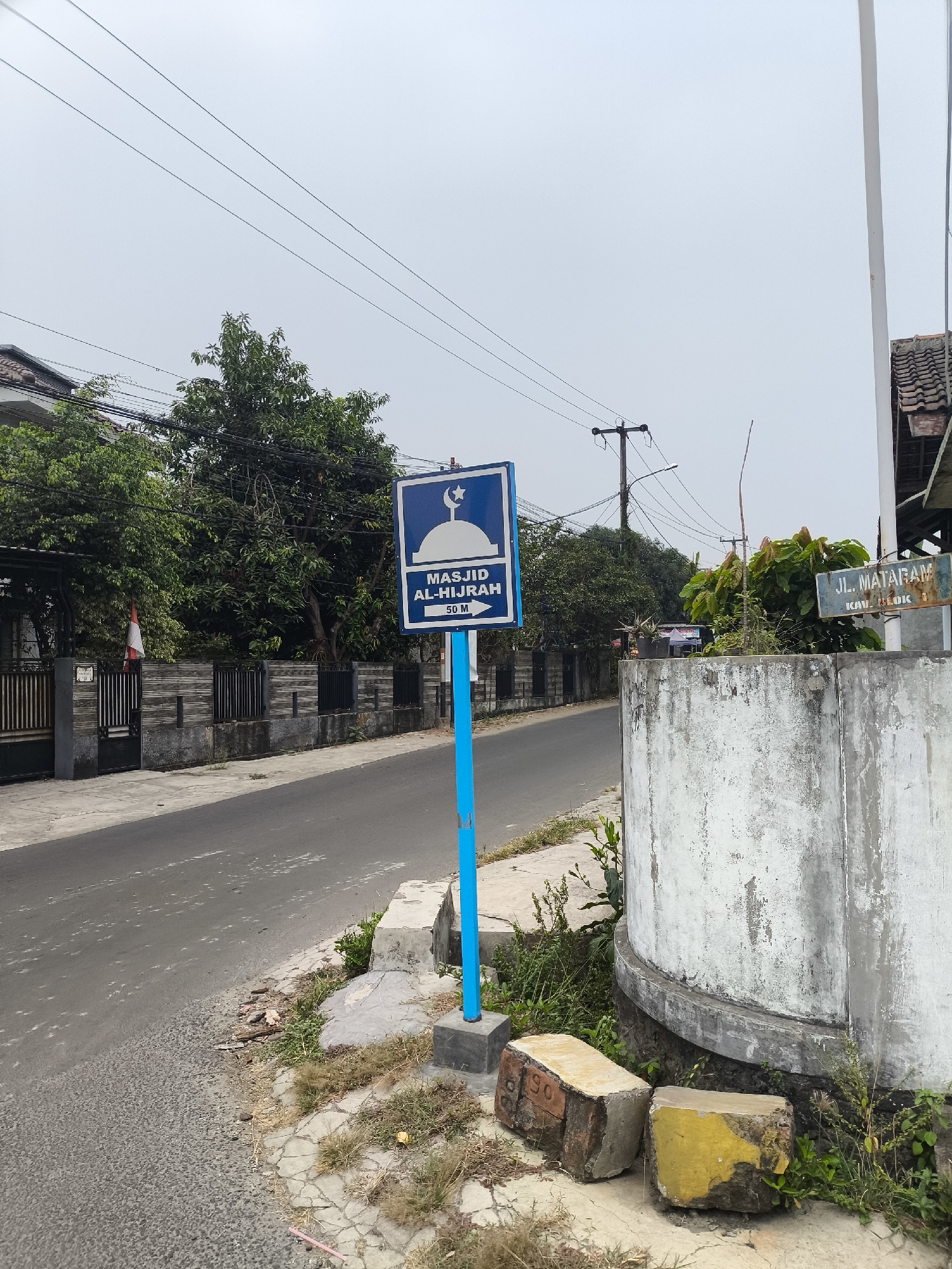The image depicts a street scene in Cilegon, Indonesia. The foreground features a blue signpost with a directional arrow pointing to the right, indicating "MASJID AL-HIJRAH 50 M". A blue sign with a mosque silhouette and crescent moon is attached to the post. To the right of the signpost is a weathered, cylindrical concrete structure with peeling white paint. Two broken bricks, one with yellow paint and the other with red markings, rest on the ground near the concrete structure. Behind this, a street curves to the left, bordered by a dark metal fence with a stone base. A red and white Indonesian flag is visible on the fence. Lush green trees line the street beyond the fence. In the background, utility poles with numerous wires are silhouetted against an overcast sky. Further to the right, another sign reads "JL. MATARAM" with smaller text underneath, indicating a street name. The environment appears to be a residential or semi-commercial area.
