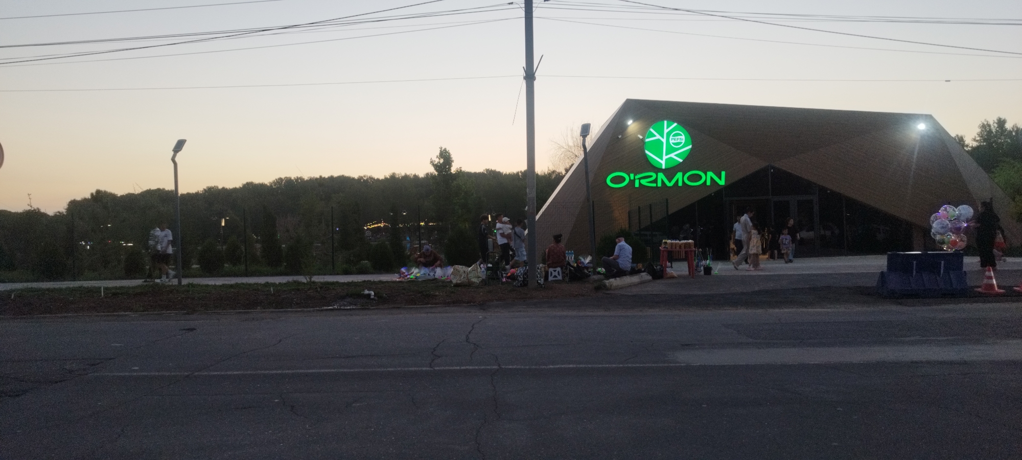 This media file captures an outdoor scene in the late afternoon or early evening, with a warm, hazy sky suggesting a clear day. A modern, angular building with a large, illuminated green logo that reads "O'RMON" is the focal point. The building appears to be a commercial establishment, possibly a restaurant or cafe, with large glass doors and windows.
In front of the building, several people are gathered. Some are sitting on the ground, seemingly resting or socializing, while others are standing or walking. There are various objects scattered around, including bags, blankets, and possibly some items for sale, such as balloons held by a person near the right side of the frame. A small red table with drinks or food is also visible.
To the left of the building, a person in athletic wear is walking on a paved path. Behind the immediate area, a dense line of trees forms a natural backdrop, with a fence running along its edge. Power lines are visible crisscrossing the sky, indicating the presence of infrastructure. The overall atmosphere suggests a relaxed, public gathering spot, possibly a park or an area adjacent to a recreational facility, where people are enjoying the end of the day. There is also a visible Pepsi logo within the larger O'RMON logo.