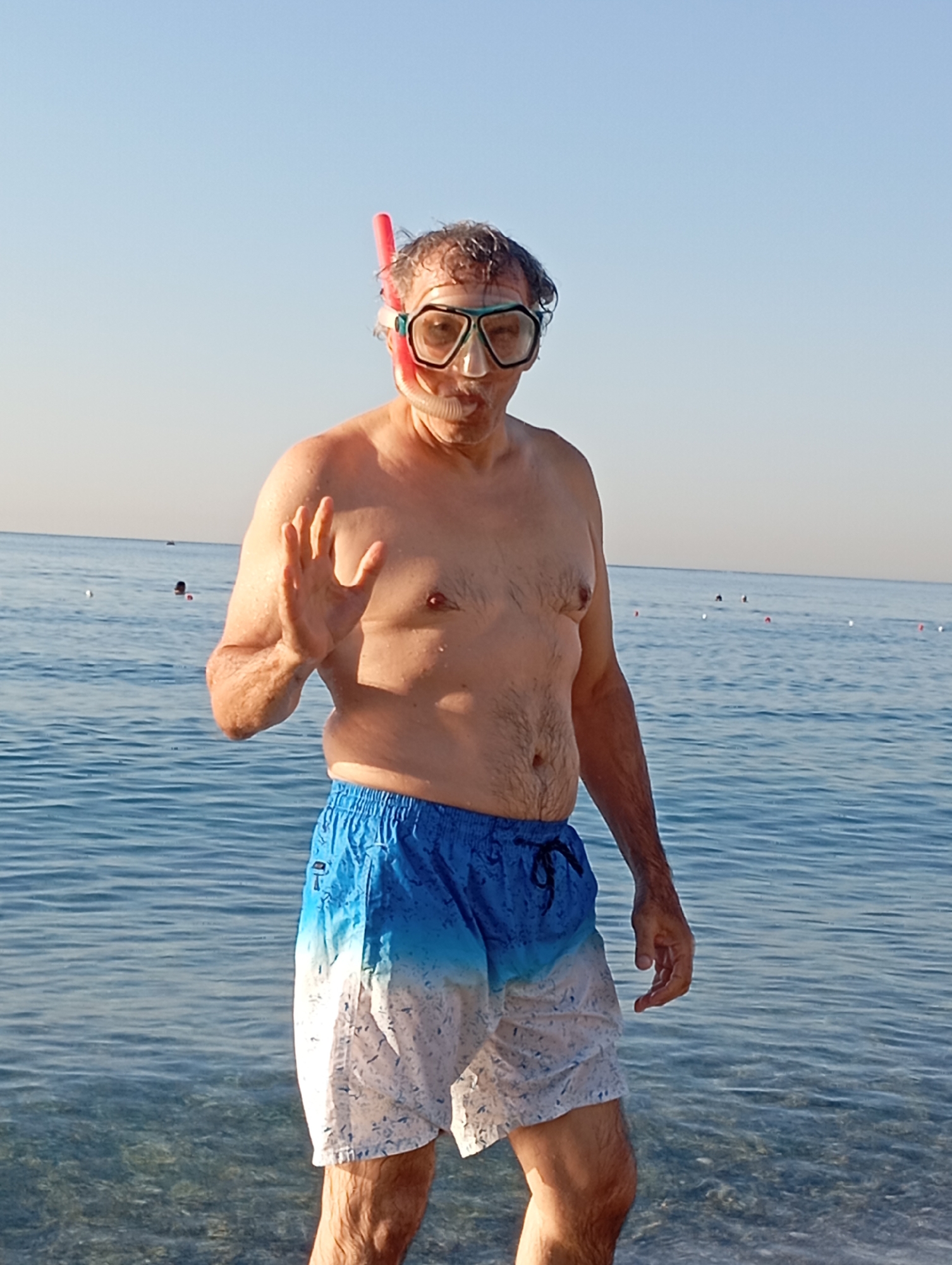 A man in his 50s stands in the shallow, clear water of a beach, wearing snorkeling gear and blue and white patterned swim trunks. The snorkel mask, with its teal frame and clear lens, covers his eyes, and a pink snorkel tube extends upwards from the mask. His hair is dark and appears wet. He is raising his left hand, palm facing outwards, in a gesture that could be a wave or a sign of stopping. The background shows a calm, blue sea under a clear, pale blue sky, indicating a sunny day. The horizon is visible, with a few indistinct figures in the water further out. The time of day appears to be late afternoon or early morning, given the soft, even lighting and the gentle hue of the sky. The man's expression, visible through the mask, seems playful or perhaps slightly surprised. The scene evokes a sense of vacation and leisure. The location is identified as Konyaaltı, Turkey, a popular beach resort area. There is no visible text in the image.