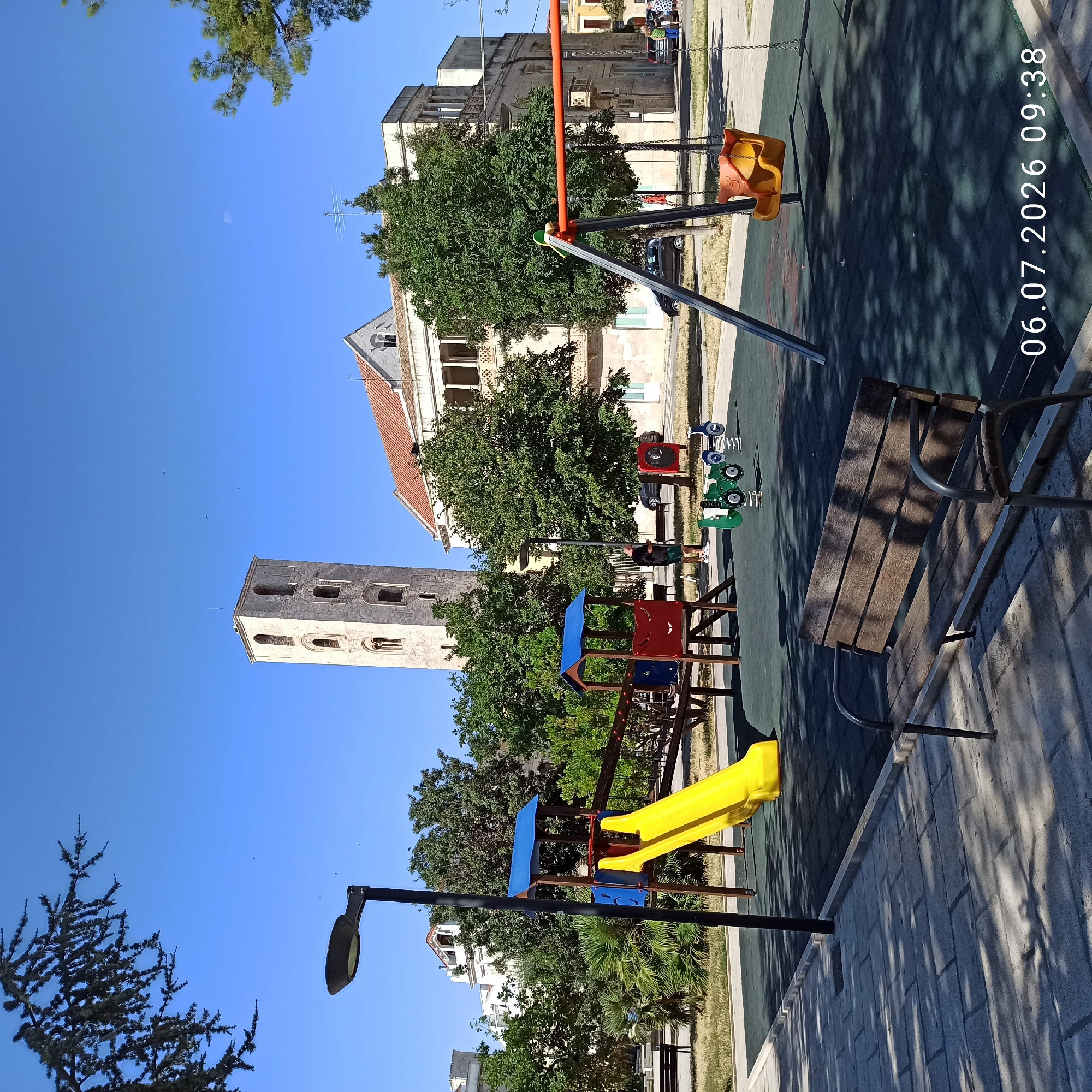 The image depicts a playground area in Ruvo di Puglia, Italy, on July 6, 2026, at 09:38. In the background, a prominent stone bell tower with arched windows stands against a clear blue sky. To the right of the tower is a building with a red-tiled roof and balconies.
The foreground is dominated by playground equipment. A yellow slide descends from a multi-colored play structure. To the right, a swing set with an orange seat is visible. A green spring rider resembling a toy tractor is also present. A wooden bench with metal legs is positioned in the lower part of the frame, casting shadows from nearby trees.
A person in dark clothing and shorts stands near the play structure, looking at their phone. Another person, partially obscured, is visible in the background on the right. Several cars are parked along the street behind the playground. The ground is a dark rubberized surface, bordered by a paved walkway with cobblestone patterns. Lush green trees and some palm trees surround the playground, indicating a public park or square. The lighting suggests it is a bright, sunny morning.