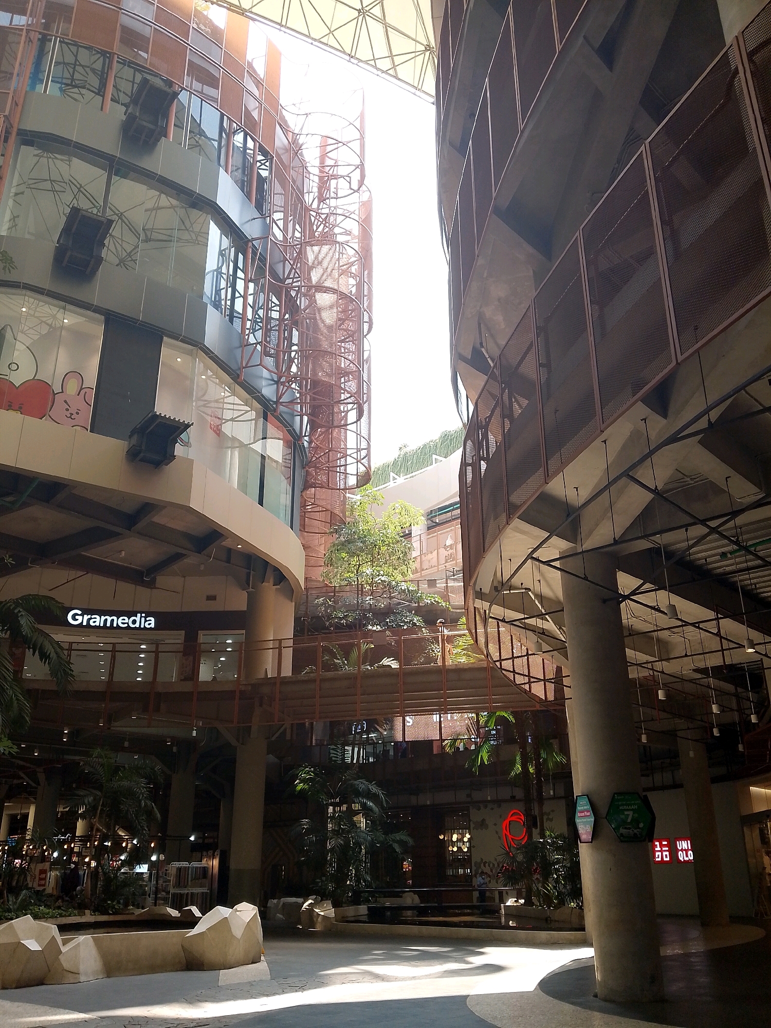 The image depicts the interior of the Trans Studio Mall in Makassar, Indonesia. The mall features a striking, multi-story architectural design with a central atrium that allows natural light to filter in from a large, transparent ceiling structure. The buildings are characterized by curved facades, glass windows, and open-air walkways with metal railings.
In the foreground, a wide, open pathway is visible, with light and shadow playing across its surface, suggesting a sunny day. The pathway is lined with abstract, light-colored stone sculptures, possibly intended as seating or decorative elements.
Various levels of the mall are visible, showcasing shops and retail spaces. One prominent sign reads "Gramedia," indicating a bookstore. Other signs and logos, such as those for "UNI QLO," are also visible, identifying retail chains.
Greenery is integrated into the mall's design, with palm trees and other tropical plants visible on different levels, creating an oasis-like atmosphere within the urban structure. A spiral staircase made of reddish-brown metal is a notable architectural feature, connecting different floors.
The overall impression is one of a modern, spacious, and well-lit indoor environment designed for shopping and recreation, incorporating elements of nature and contemporary design. No specific individuals are clearly identifiable or engaged in any particular actions within the frame.