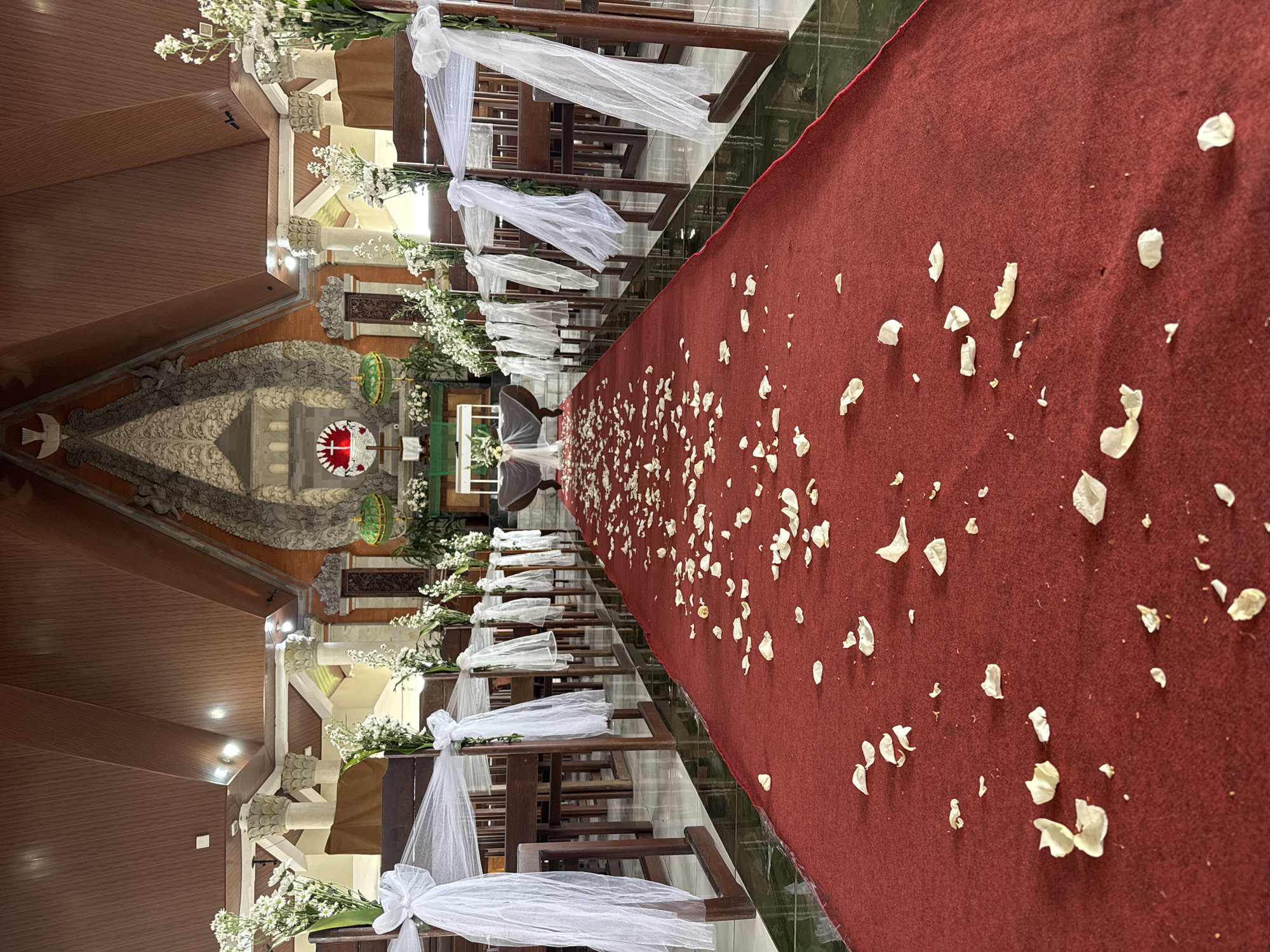 The image depicts the interior of a wedding venue in Kuta Selatan, Indonesia. A red carpet runner is laid out, leading towards an altar area. The carpet is scattered with white flower petals. Wooden pews, decorated with white tulle and flowers, line either side of the carpet. The altar features a cross and what appears to be decorative greenery. The ceiling is made of wood, with exposed beams and lighting fixtures. The architecture suggests a church or chapel setting, possibly with Balinese influences given the location. No people are visible in the image.