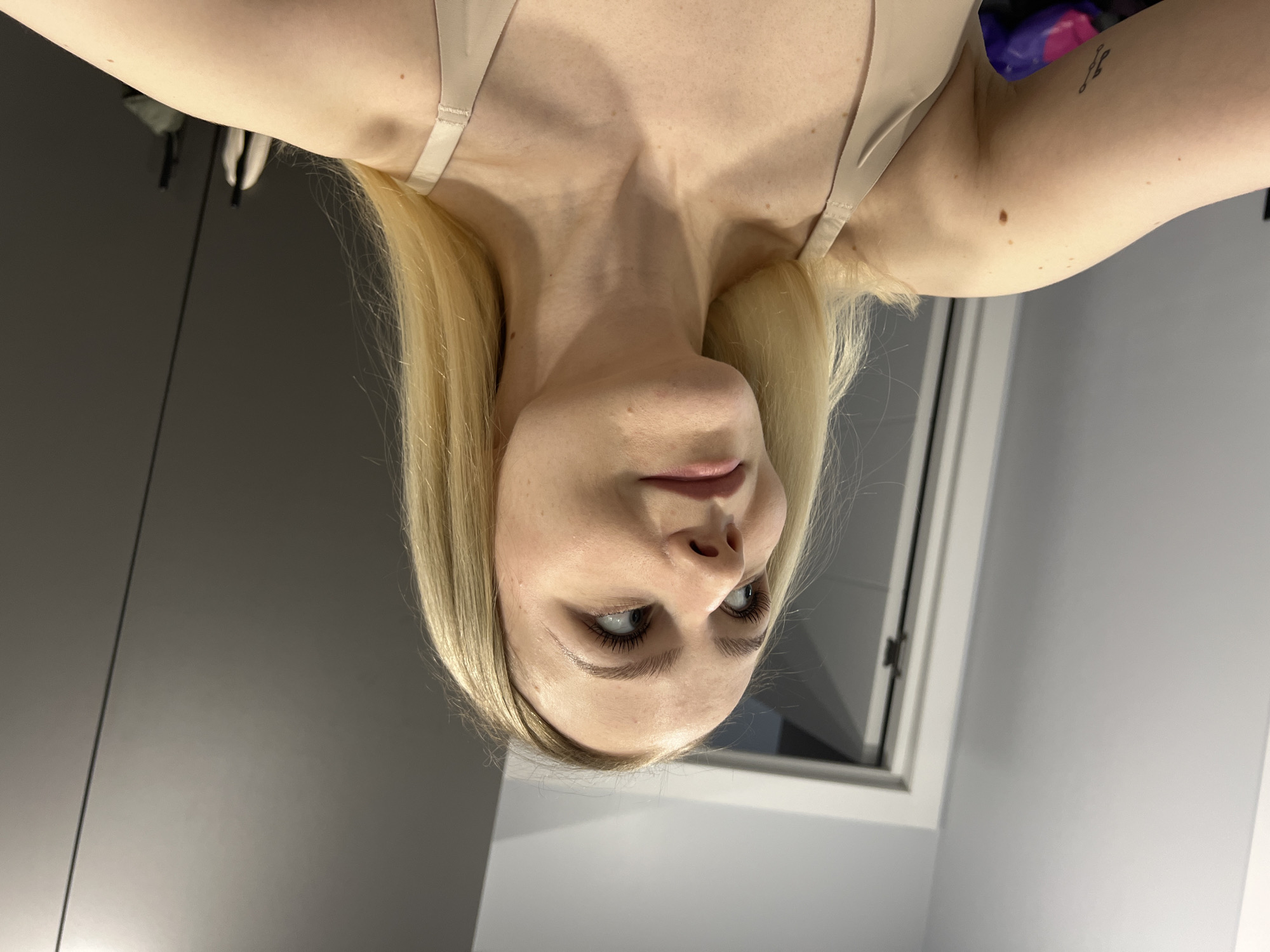 A fair-skinned individual with blonde hair is depicted from an upside-down perspective. The person is wearing a light-colored bra or tank top. The background includes a gray wall, a window, and a glimpse of some items in the upper right corner. A tattoo is visible on the person's upper arm.