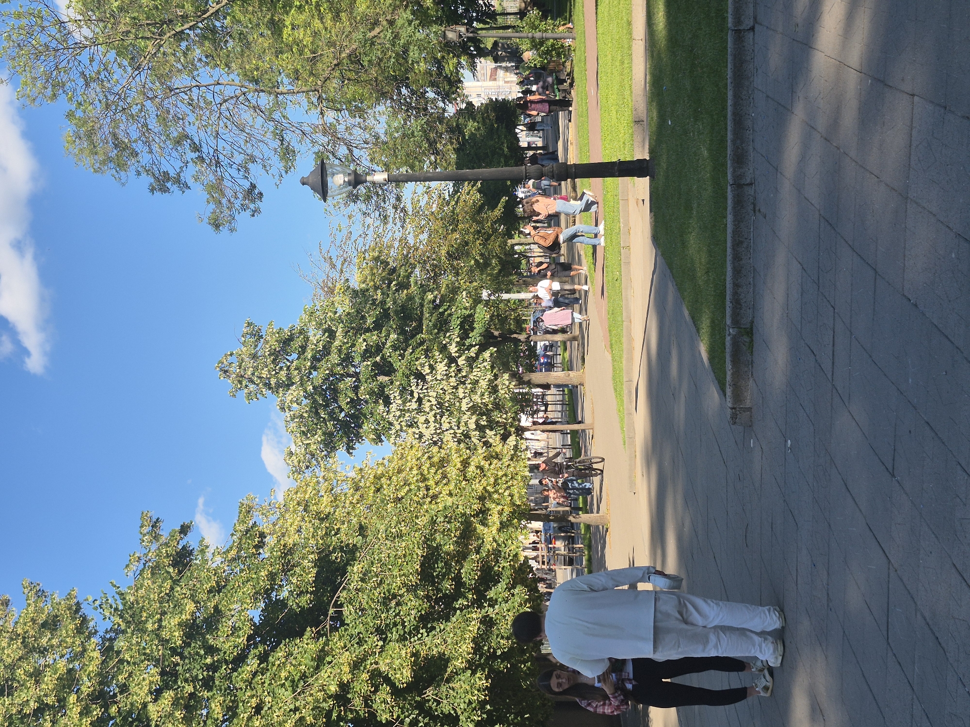 The image depicts a sunny day in a park in Chișinău, Moldova. In the foreground, two individuals are standing on a paved walkway. One is a person wearing a white outfit, possibly a uniform, and the other is a woman wearing a plaid shirt and black pants. Behind them, a paved path winds through a park with lush green trees and grass. Several other people are visible walking and cycling in the background. A lamppost stands prominently in the middle ground. The sky is clear blue with a few scattered clouds. The overall atmosphere suggests a pleasant day in a public urban space.