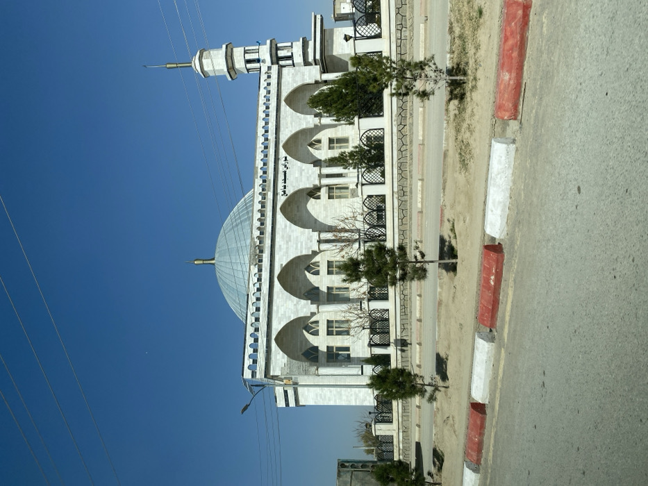 The image displays the Shurai Grand Mosque (Masjid Jame'e Shura'i) in Kandahar, Afghanistan, under a clear blue sky. The mosque is a large, light-colored building with a prominent, ribbed, light-blue dome topped by a golden finial. A multi-tiered minaret, also featuring a golden finial, stands on the right side of the main structure. The mosque's facade incorporates several arched recesses above rectangular windows. Arabic script, transliterating to "Shurai Grand Mosque," is inscribed above the central arches. The mosque property is enclosed by a black, ornate metal fence, behind which several small evergreen trees and some bare deciduous trees are visible. In the foreground, an asphalt road is present, bordered by a curb composed of alternating red and white painted blocks. A narrow dirt median containing several young evergreen trees separates the curb from the mosque grounds. Overhead, multiple power lines stretch across the upper portion of the frame, with a streetlamp visible on the far left. No individuals are present in the scene.