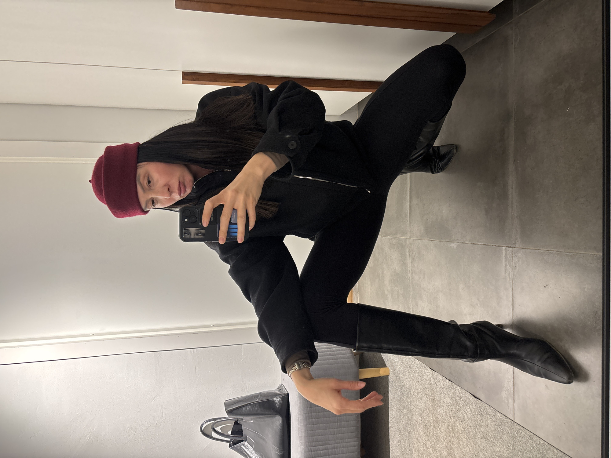 A woman is taking a selfie in a dressing room. She is wearing a maroon beanie, a black jacket, black leggings, and black leather boots. She is holding a smartphone in her hands. A black handbag is visible on a gray bench. The dressing room has a gray tile floor and white walls. A wooden strip runs horizontally along one of the walls. The location appears to be an indoor retail space, not necessarily in Chaikovsky, Russia, as that detail cannot be determined from the image.