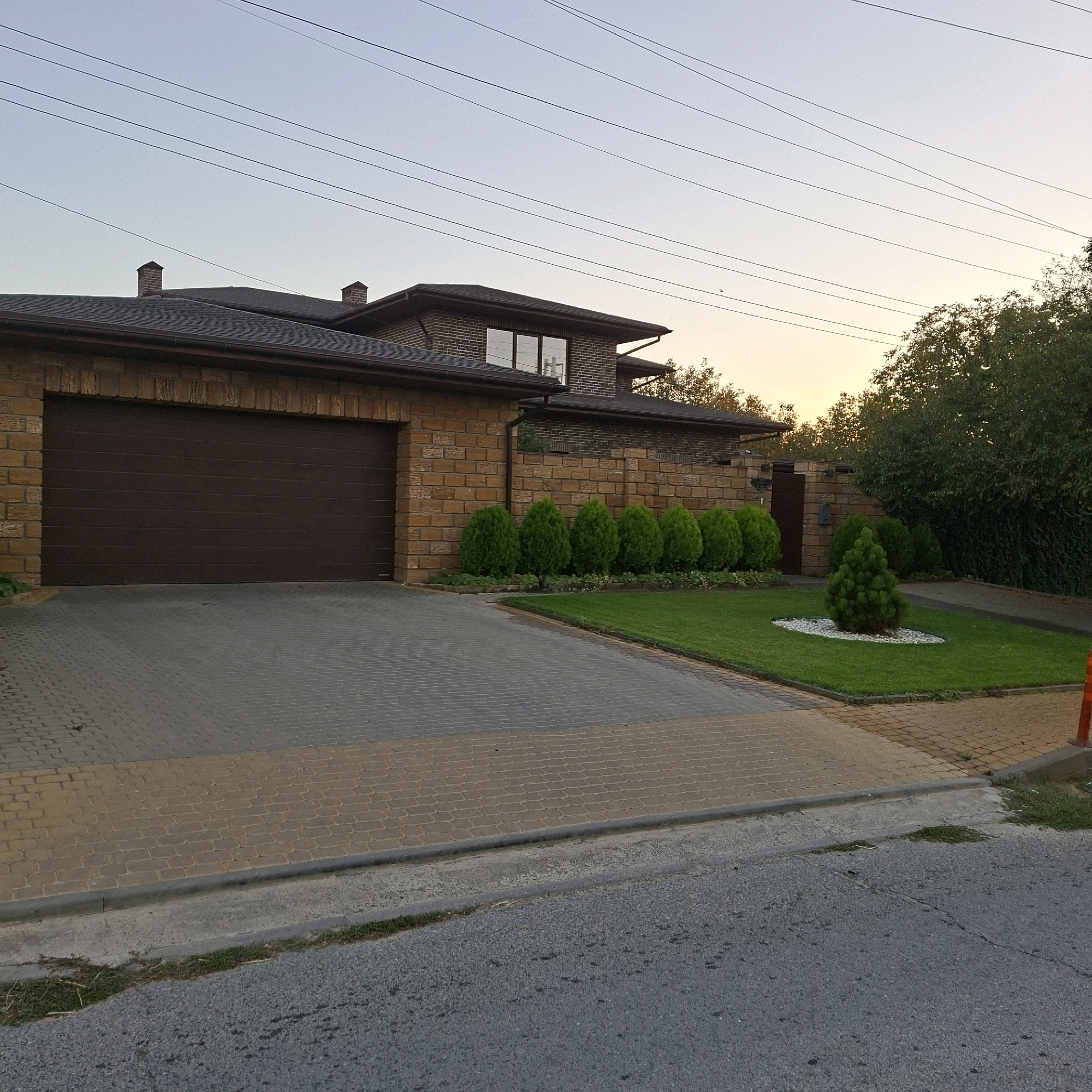 The image captures a residential property in Anapskaya, Russia, likely taken during the late afternoon or early evening given the soft, warm light. The scene is dominated by a large, modern house constructed with light brown brick. A dark brown, horizontal panel garage door is prominent on the left side of the house. The driveway leading to the garage and continuing in front of the house is paved with interlocking concrete pavers, a mix of gray and light brown.
The house features a dark roof and several windows, some reflecting the sky. A brick wall, also made of the same light brown brick, surrounds part of the property. In front of this wall, a neatly manicured lawn is bordered by a line of evenly spaced, cone-shaped evergreen bushes. Further to the right, another small, conical evergreen tree stands on a circular bed of white gravel. Beyond the manicured lawn, dense green trees form a natural backdrop, with a sliver of the sky showing a gradient of light blue and pale yellow, suggesting sunset. Power lines are visible crisscrossing the sky. A small, orange traffic cone is visible at the very edge of the frame on the right. The foreground shows a section of asphalt road with a concrete curb. There are no people visible in the image.