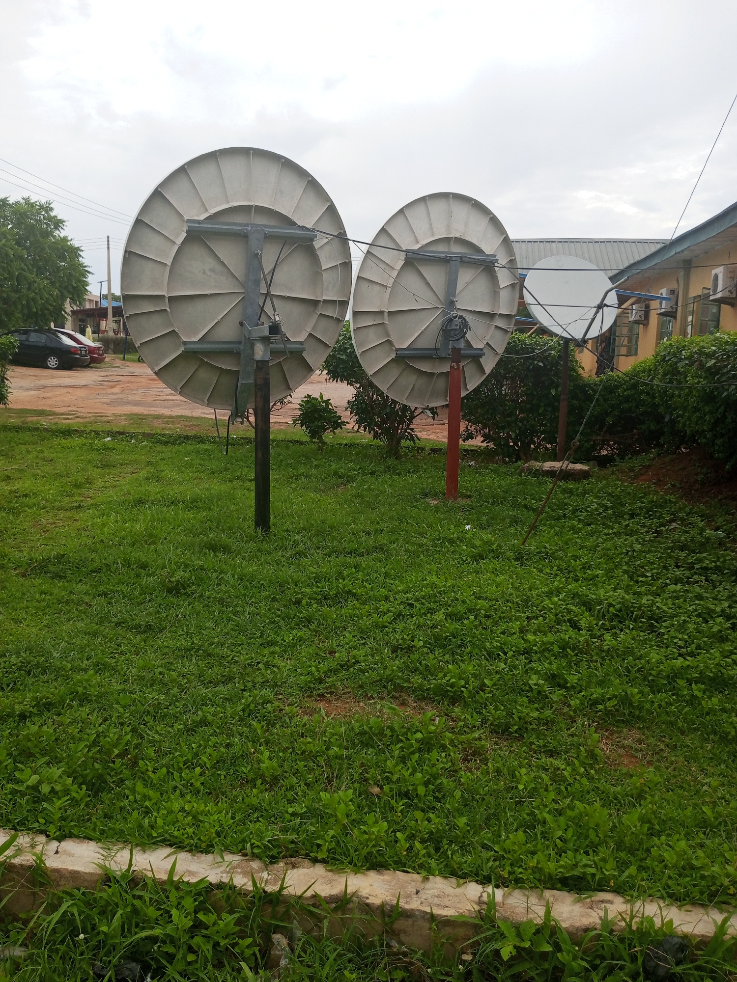 This image captures a scene in Jalingo, Nigeria, likely during the daytime. The weather appears overcast, contributing to a muted, diffused light. The primary subjects are three large satellite dishes positioned in a grassy area. Two of the dishes are prominently featured in the foreground, facing away from the camera. They are mounted on poles, with one pole being a dark wood or metal and the other a reddish-brown. The dishes themselves are a light, neutral color, with visible structural supports and segmented surfaces. Behind these, a third, smaller satellite dish is partially visible, also mounted on a pole.
The setting includes a well-maintained lawn with a variety of green vegetation, suggesting an outdoor area, possibly near a building. In the background, a building with a light-colored facade and a dark-pitched roof is visible. Parts of its exterior, including windows and air conditioning units, can be discerned. To the left of the frame, several parked cars are visible on a dirt or gravel surface, indicating a possible parking area or access road. Trees and dense green bushes form a backdrop to the satellite dishes, further defining the outdoor environment.
There is no clear activity or event taking place in the image; it appears to be a static shot of the installation. The overall mood is neutral, with the overcast sky and focus on the equipment creating a functional rather than emotive atmosphere. No visible text is present in the image. The scene is likely a communication hub or a place where satellite reception is important, given the presence of multiple dishes.