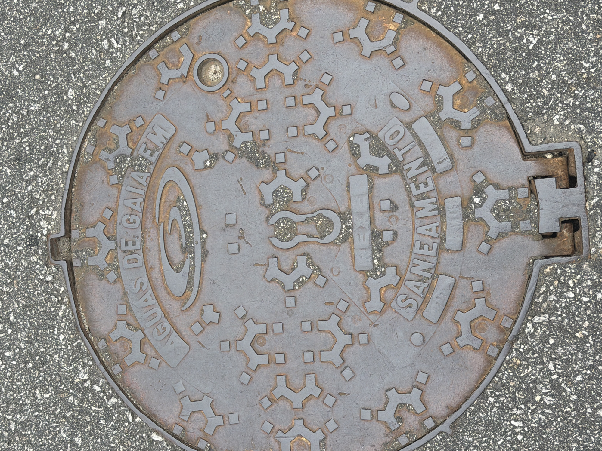 The image shows a circular, weathered metal manhole cover embedded in asphalt pavement. The manhole cover features embossed text that reads "ÁGUAS DE GAIA EM SANEAMENTO" in an arc around the outer edge. A central logo, possibly depicting water flow, is also visible. The cover is decorated with a geometric pattern of interconnected shapes resembling gears and small squares. The surrounding asphalt appears to be a standard city street surface with visible aggregate. The lighting suggests it is daytime. The manhole cover is a utility cover, likely for the water and sewage system in Vila Nova de Gaia, Portugal.
