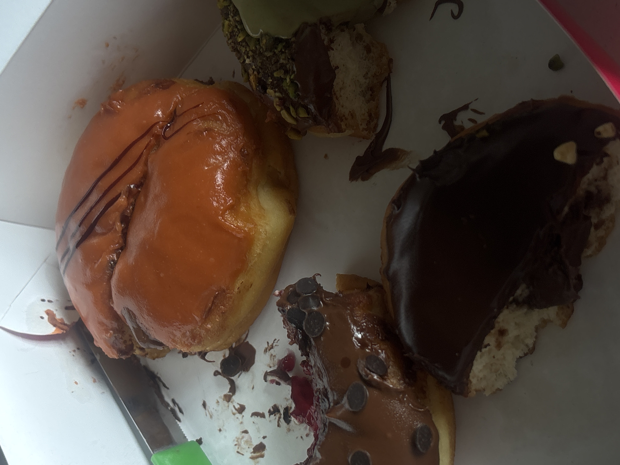 A high-angle, close-up shot reveals a partially eaten box of donuts. One donut has orange frosting with chocolate drizzle. Another has a chocolate glaze, revealing the white interior. A third donut has a light brown glaze with small chocolate chips. A fourth donut has light green sprinkles around the top. The donuts sit inside a white cardboard box. A knife with a green handle lays to the left of the orange donut. The location is Yerevan, Armenia.