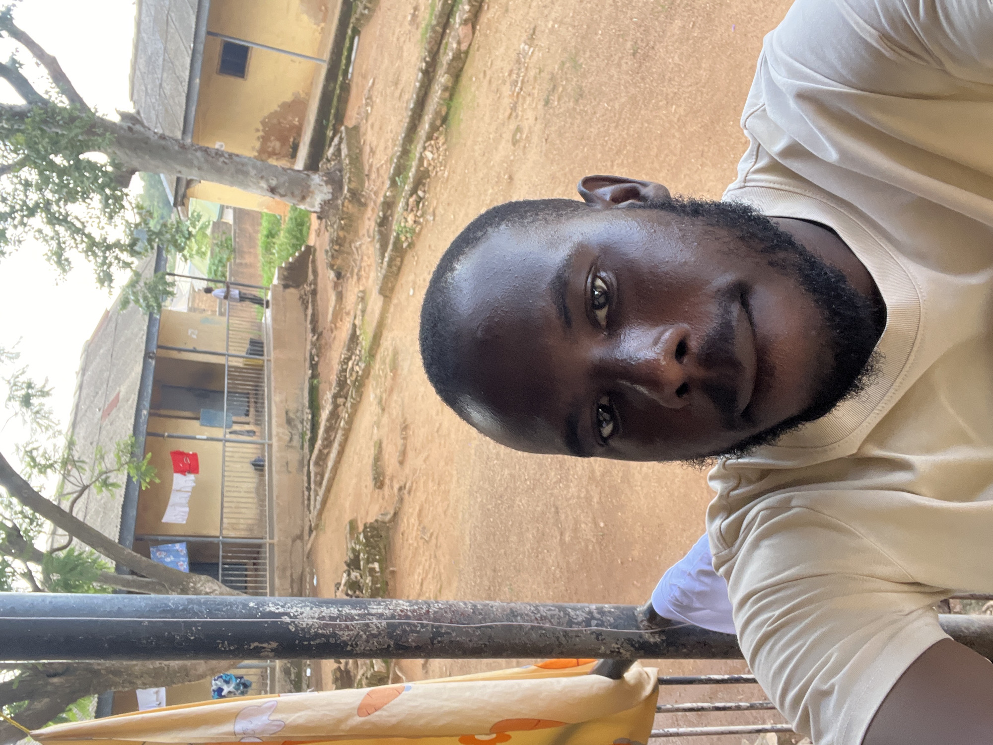 A medium shot selfie is taken by a man with dark skin and a beard, wearing a light-colored t-shirt. He is looking directly at the camera. Behind him, a pole supports a yellow cloth with an orange pattern. Beyond the pole, there are two yellow buildings with tin roofs and dark windows. Clothes are hanging on a line in front of one of the buildings. The ground is covered in reddish-brown dirt. A large tree with visible roots stands between the buildings. In the background, there are other structures and greenery, suggesting an outdoor setting in a residential area. The overall scene appears to be in a warm climate, likely during daylight hours. The location is identified as the city of Jos, Nigeria.