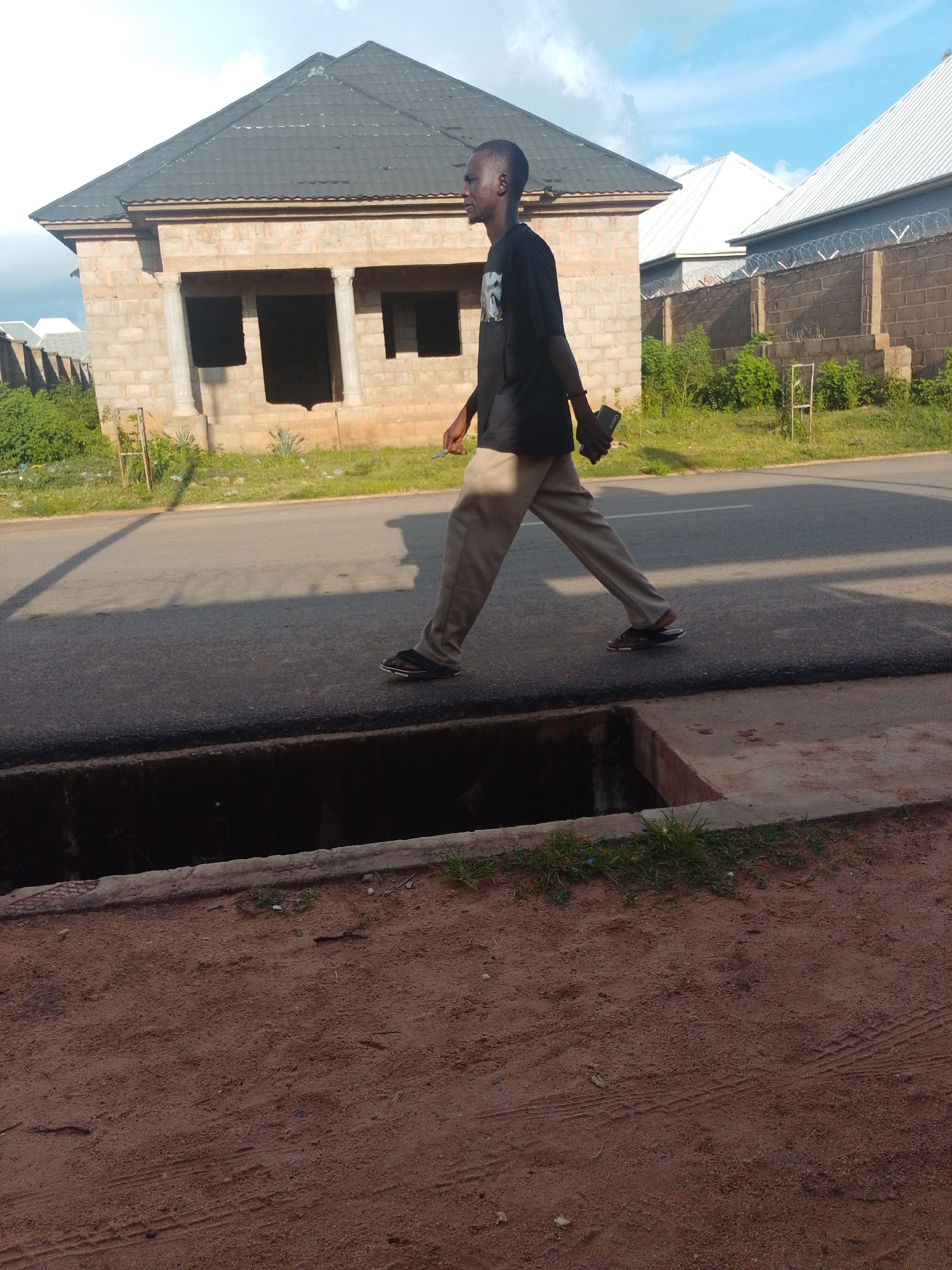 A side profile, full-body shot shows a young Black man in motion walking across a paved road in front of a partially constructed building. The man is wearing a black t-shirt and tan pants, and he has dark hair. He is carrying a dark-colored object in his right hand, possibly a phone. His body is angled to the right, suggesting he is walking towards the right side of the frame. The road has a slight sheen, indicating it may have been recently paved. In the foreground, a drainage ditch is visible, its concrete edges worn and covered in sparse grass. Behind the man, the building under construction features brick walls, visible pillars, and dark, empty window and door openings. Beyond the building, more structures with light-colored roofs and brick walls with barbed wire fencing are present. The sky above is a pale blue with scattered white clouds. The ground on the right side of the frame appears to be dirt or sand with tire tracks.