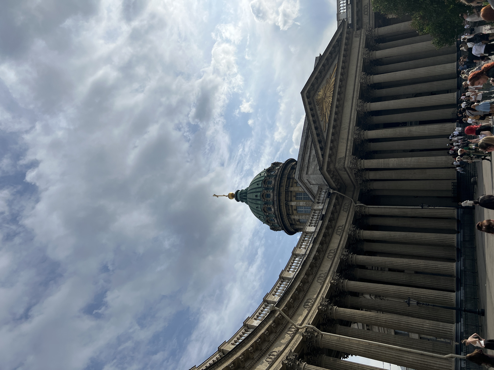 This is a low-angle, vertical shot of a large, classical building with a dome and numerous columns, set against a cloudy sky. The building's facade features intricate architectural details, including ornate carvings and a golden sunburst emblem. A green dome with a golden spire rises from the structure.
A crowd of people is gathered in front of the building, viewed from the side. They appear to be looking at or interacting with the building, or perhaps a service is taking place. The people are dressed in casual summer clothing.
The time of day appears to be daytime, indicated by the light and shadows. The weather is overcast with some breaks of blue sky, suggesting a variable or partly cloudy day. The architectural style and the presence of the dome strongly suggest this is a significant religious or governmental building. Given the provided location context of Saint Petersburg, Russia, this is likely Kazan Cathedral. The overall mood is one of respectful observation or attendance. No visible text is present in the image.