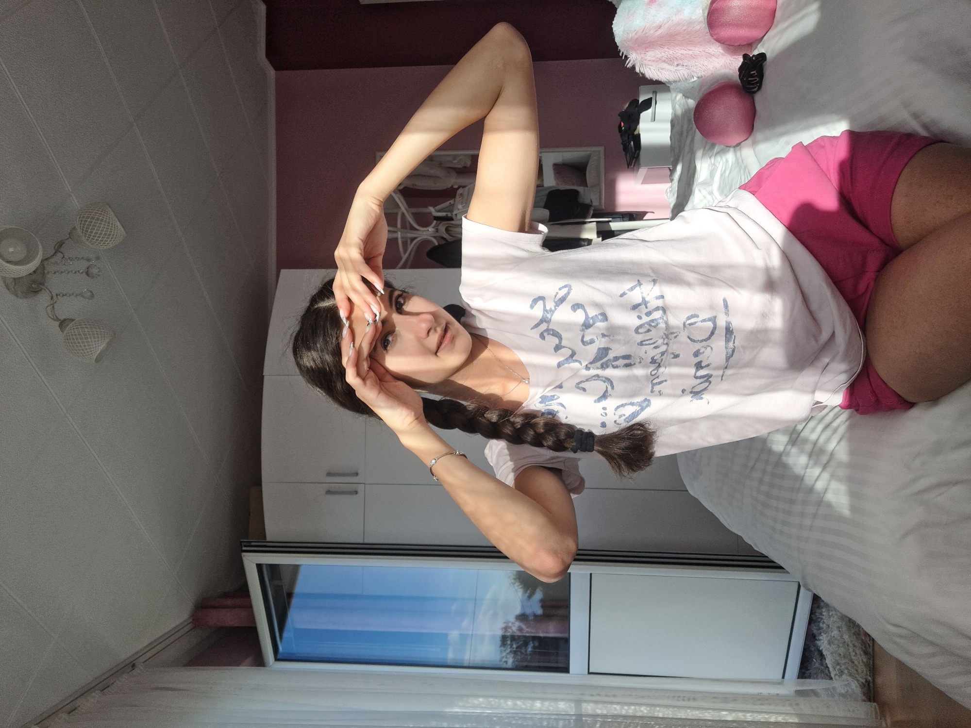 A young woman with long, dark hair in a braid is lying on a white bed with striped bedding. She is wearing a light-colored t-shirt with a graphic print and pink shorts. Her left arm is raised above her head, and her right hand is near her face. She is looking at the camera. Behind her is a white cabinet with a window, and in the background, there is a pink wall and a ceiling with a light fixture. There is also a large stuffed animal on the bed near her head.
