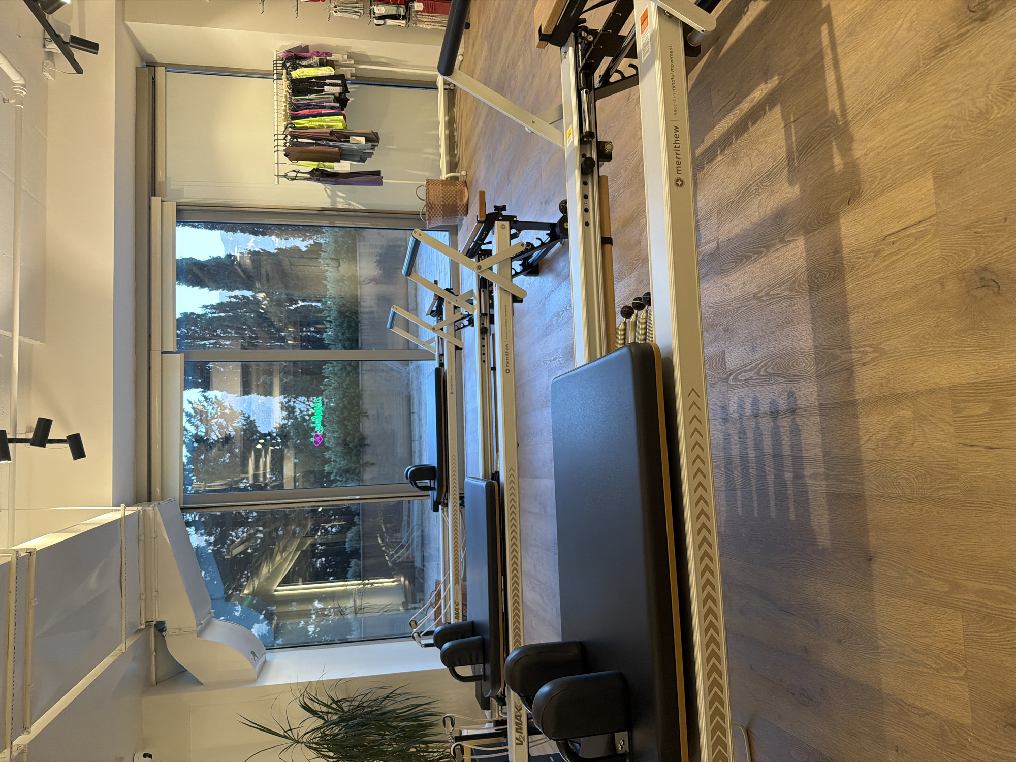 The image depicts the interior of a pilates studio, likely located in Boreti, Montenegro, as indicated by a brand name on the equipment. The studio features multiple pilates reformers arranged in parallel. These reformers are predominantly white with black padded surfaces and headrests. The flooring is a warm-toned wood laminate, and natural light streams in from a large window that offers a view of trees and distant hills.
In the background, a clothing rack displays various athletic wear items, suggesting the studio may also sell apparel. A woven basket sits near the window. The overall ambiance is clean, modern, and serene, with shadows cast on the floor from the studio equipment and overhead lighting fixtures. There are no people visible in the scene. The brand "merrithew" is prominently displayed on the reformers, along with the tagline "leaders in mindful movement." The window also features an illuminated sign with a light green cursive font, partially visible in the reflection.