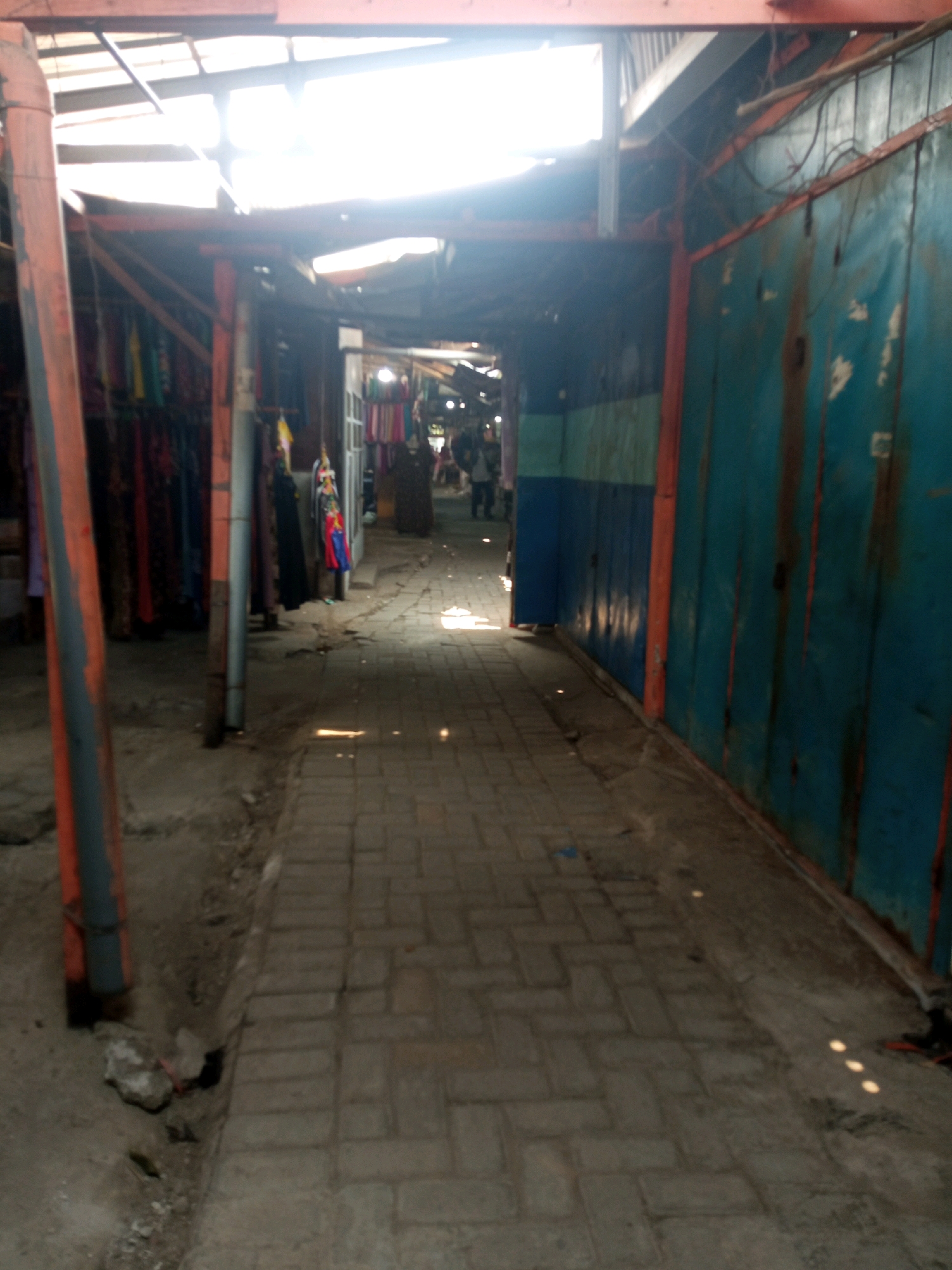The image depicts an outdoor market alleyway in Sambong, Indonesia. The alley is paved with brickwork and is lined with stalls on both sides. The left side of the alley features stalls displaying various colorful fabrics and clothing items, hung on what appear to be wooden or metal racks. These stalls are supported by orange and gray poles. The right side of the alley is dominated by large, teal-colored metal panels, suggesting the presence of enclosed shops or booths. Orange metal beams run vertically along these panels. The alley is covered by a translucent roof structure, allowing bright sunlight to filter through, creating stark contrasts between illuminated and shadowed areas on the pavement. In the background, a few people are visible walking through the alley, further indicating the active nature of the market. The overall scene suggests a typical bustling local market environment.