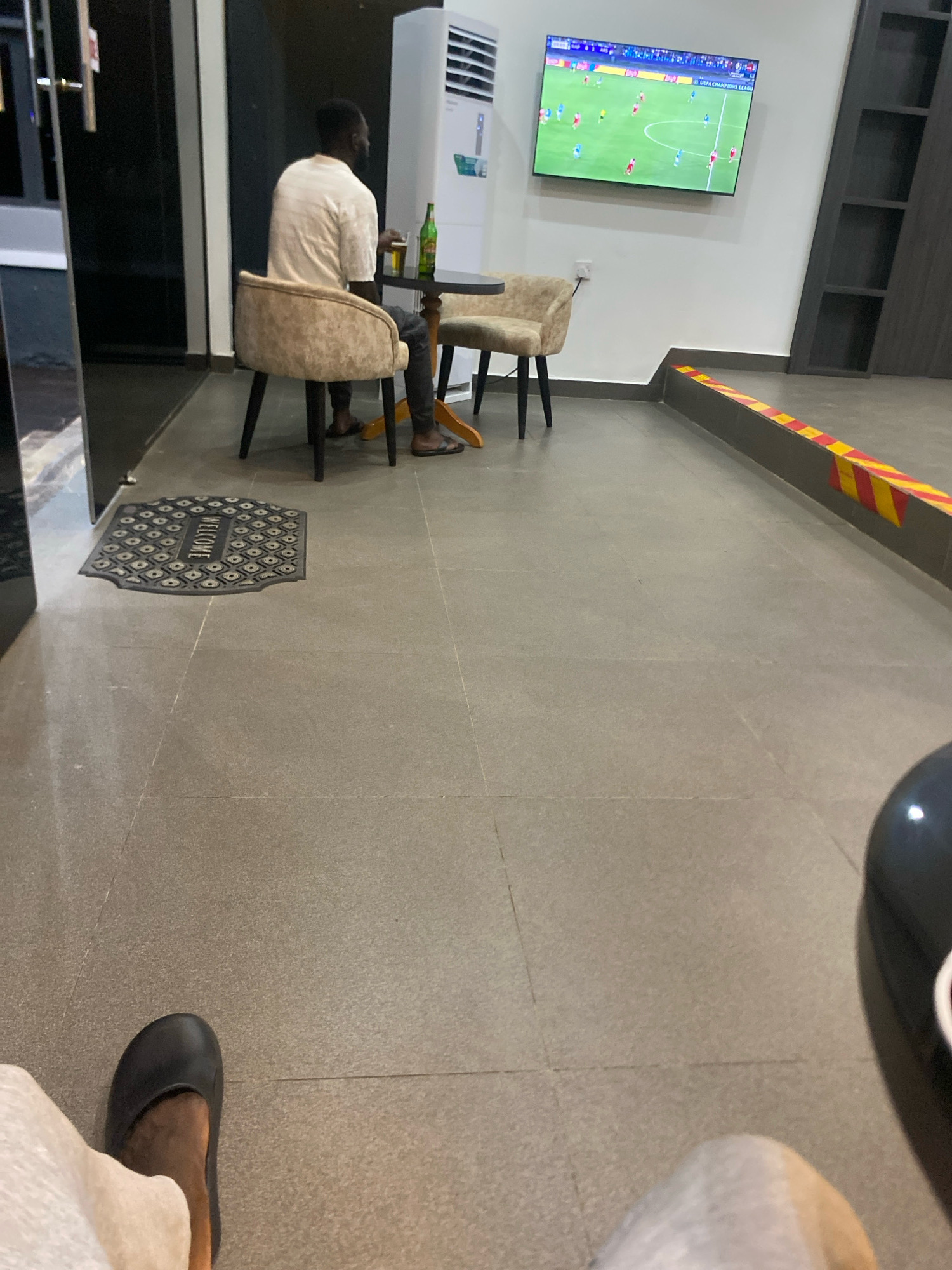 In Barden Yana, Nigeria, a person is seated at a table, facing away from the camera, with a drink and a bottle of beverage. A television screen on the wall displays a soccer game. The environment appears to be a lounge or waiting area with tiled flooring and a welcome mat.