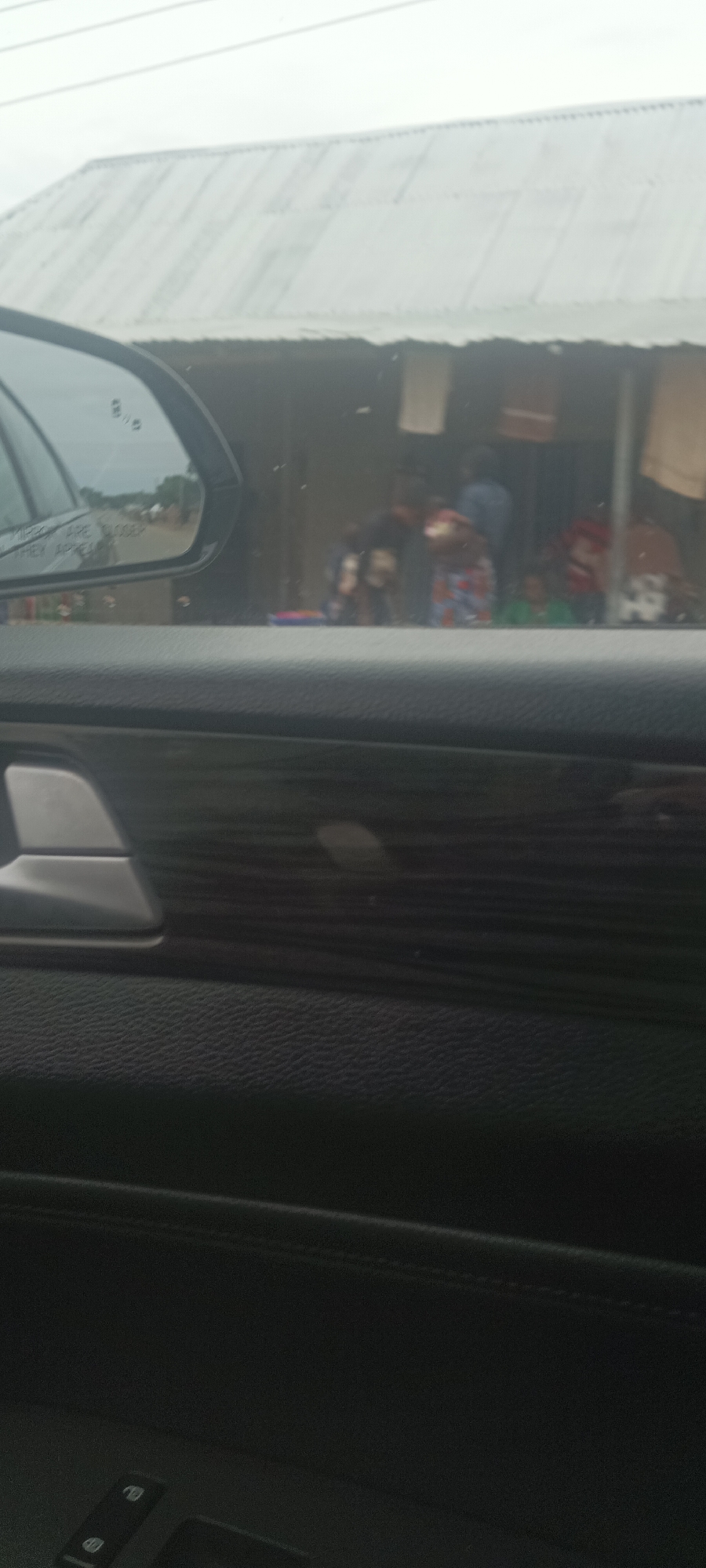 The image, likely taken from inside a car, shows a view through the window. Reflected in the car's side mirror is a road with vegetation. The background features a building with a corrugated metal roof. Several people can be seen in front of the building, and clothes appear to be hanging in front of it. The photo was taken during daylight. The city is Gwadanye, Nigeria.