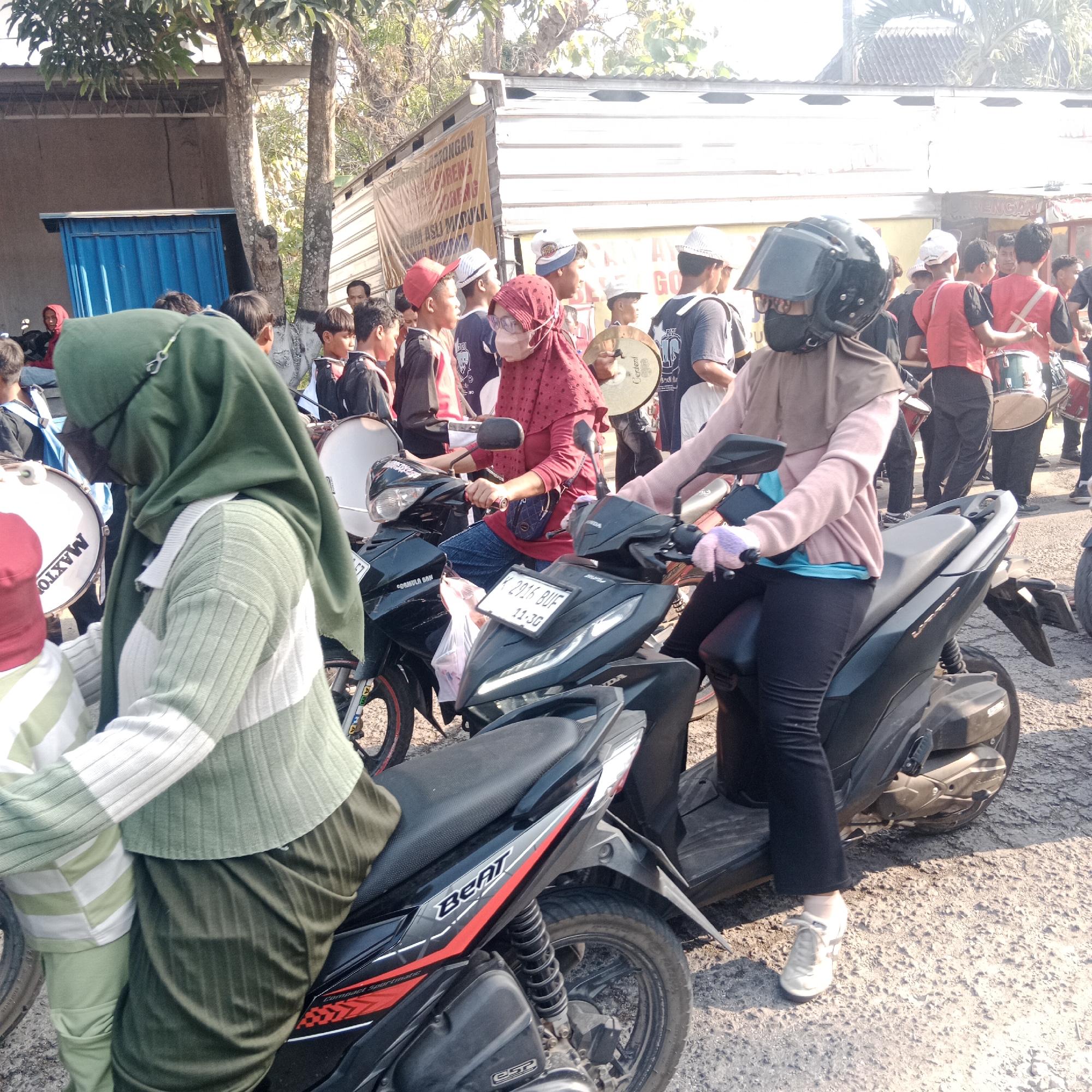 In a street scene in Tegalrejo, Indonesia, a group of people, some of whom appear to be part of a marching band, are gathered. Two women are prominently seated on black motorcycles in the foreground. The woman on the left wears a green headscarf and a striped long-sleeved shirt in green and white. The woman on the right wears a brown headscarf and a pink long-sleeved shirt, along with black pants and white sneakers. She is wearing a black helmet with a clear visor and appears to be holding something pink, possibly gloves.
Behind them, several individuals, mostly young men, are visible. Many are wearing red vests over dark shirts or jackets, and some are carrying musical instruments such as drums and cymbals. One individual in the background is holding a cymbal. The environment suggests an outdoor event, possibly a parade or a procession, with buildings and trees visible in the background. The ground is covered in gravel and dirt. A blue structure, possibly a shed or a booth, is visible to the left. The lighting indicates it is daytime.