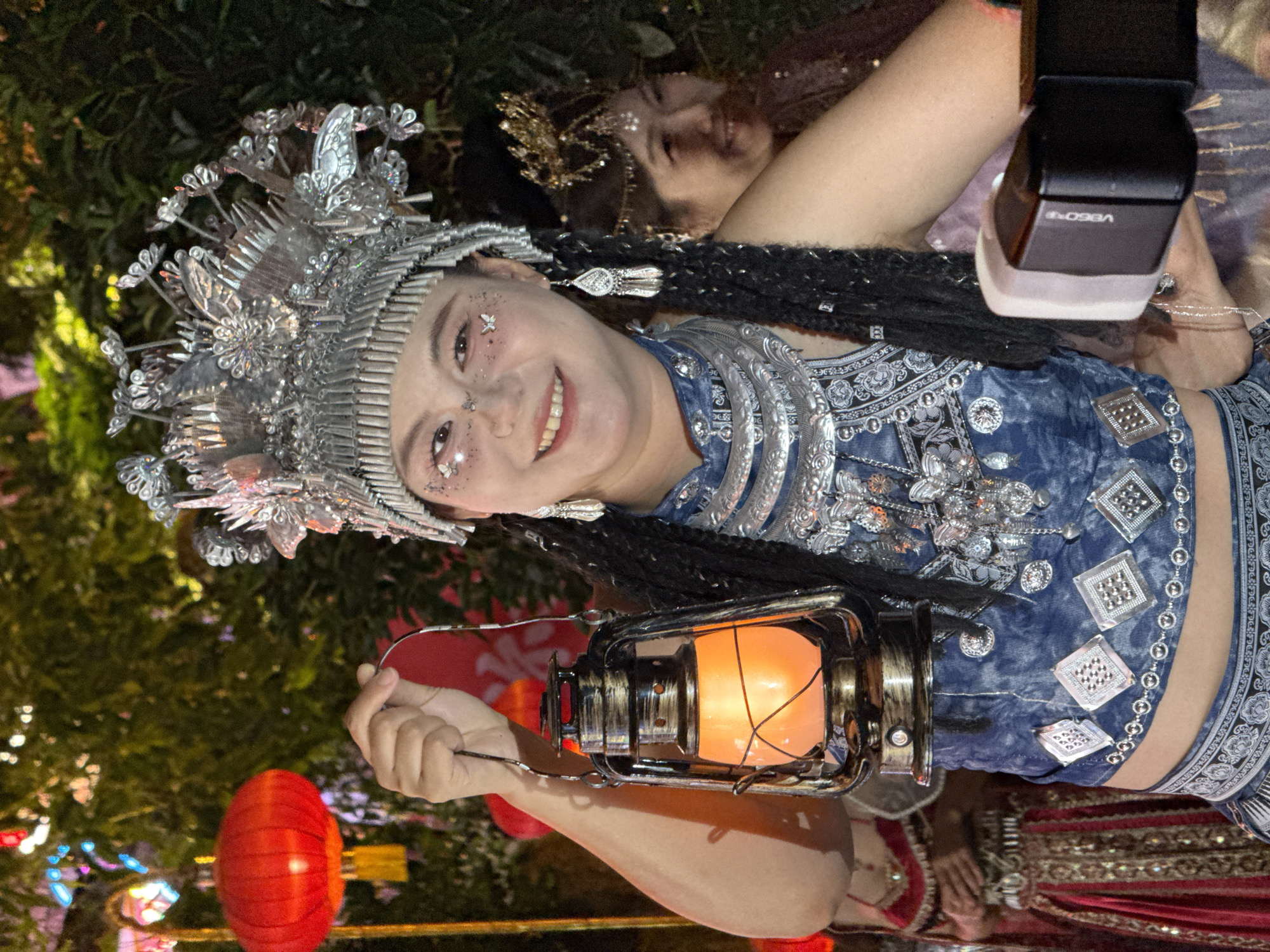 A medium, indoor shot shows a smiling woman holding a lantern, while standing alongside another woman.
The woman is holding an old-fashioned looking lantern with an orange light. She is wearing a shiny, silver headpiece. Her dark hair is in long braids. She is also wearing a blue, sleeveless shirt or top with silver embellishments. The other woman is wearing a gold headpiece, and a purple top. A camera is pointing at the women. There are Chinese lanterns hanging behind them. The location appears to be Yuzhong District, China.