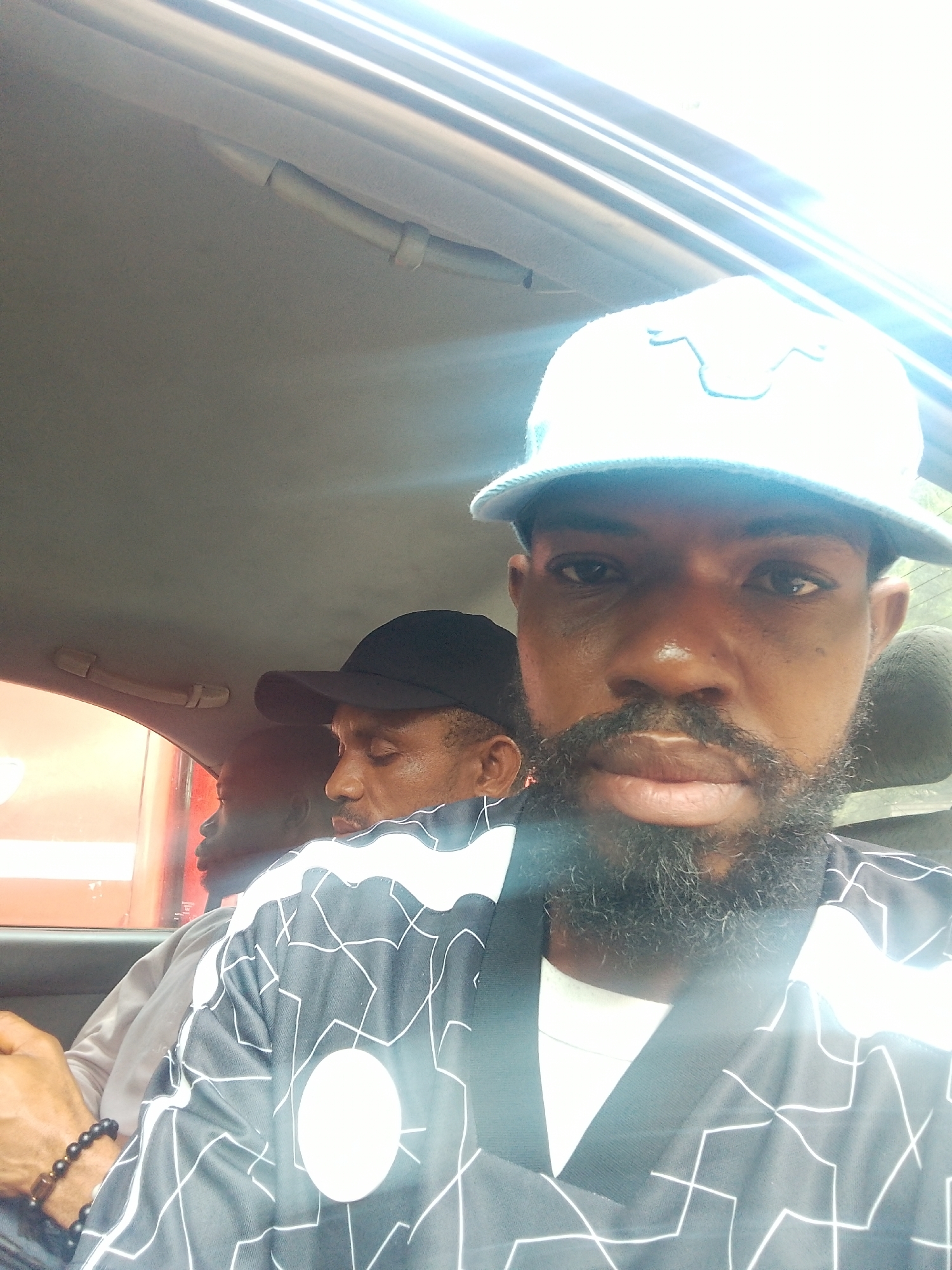 The media file shows three men inside a vehicle, likely a car, with one man in the foreground taking a selfie. The man in the foreground is wearing a light blue baseball cap and a black and white patterned shirt over a white t-shirt. He has a beard and is looking directly at the camera. The man behind him, to his left, is wearing a black baseball cap and is looking to the side. The third man is partially visible behind the man with the black cap, also looking to the side. The interior of the car is visible, including the ceiling and a grab handle. The lighting suggests it is daytime, possibly with bright sunlight coming through the windows. The overall setting appears to be casual, with the men likely in transit. The context provided indicates the location as Iju, Nigeria. There is no visible text in the image.