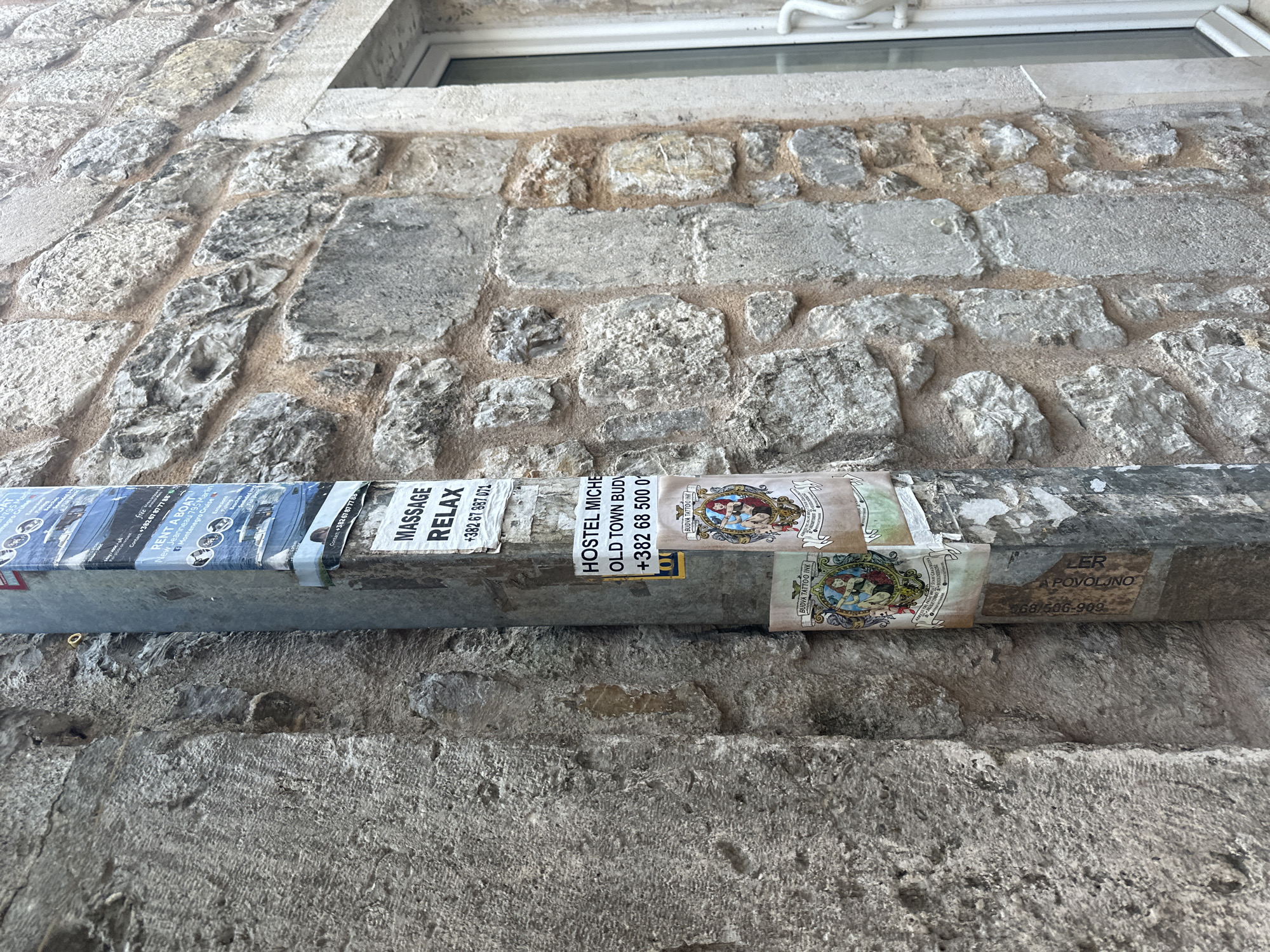 The image shows a metal bar attached to a stone wall in Budva, Montenegro. The bar is covered with several advertisements, including one for boat rentals ("RENT A BOAT Montenegro Cruise"), a massage service ("MASSAGE RELAX"), a hostel ("HOSTEL MICHE"), and a tattoo parlor ("BUDVA TATTOO INK"). Phone numbers are included on some of the ads. The wall is made of rough-hewn stones with visible mortar. Part of a window frame is visible at the top of the image.