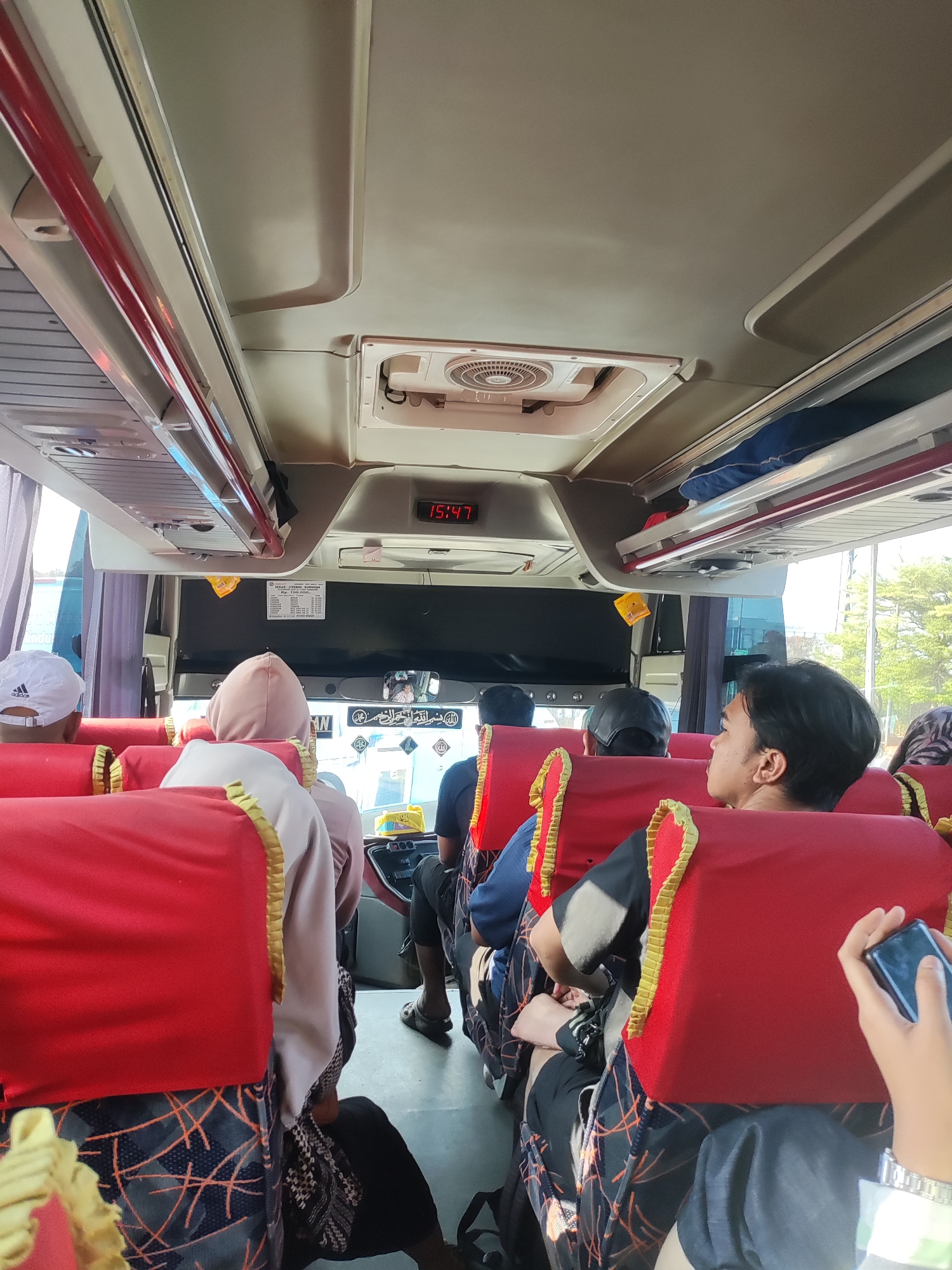 The image shows the interior of a bus, likely in Cikampek, Indonesia, with several passengers seated. The bus has red upholstered seats with gold trim. Above the seats, there is overhead storage. A digital clock on the ceiling displays the time as "15:47". In the foreground, a hand holds a smartphone. Passengers are seen from behind, some wearing headscarves. The environment outside the bus windows appears to be bright with some greenery, suggesting daytime.