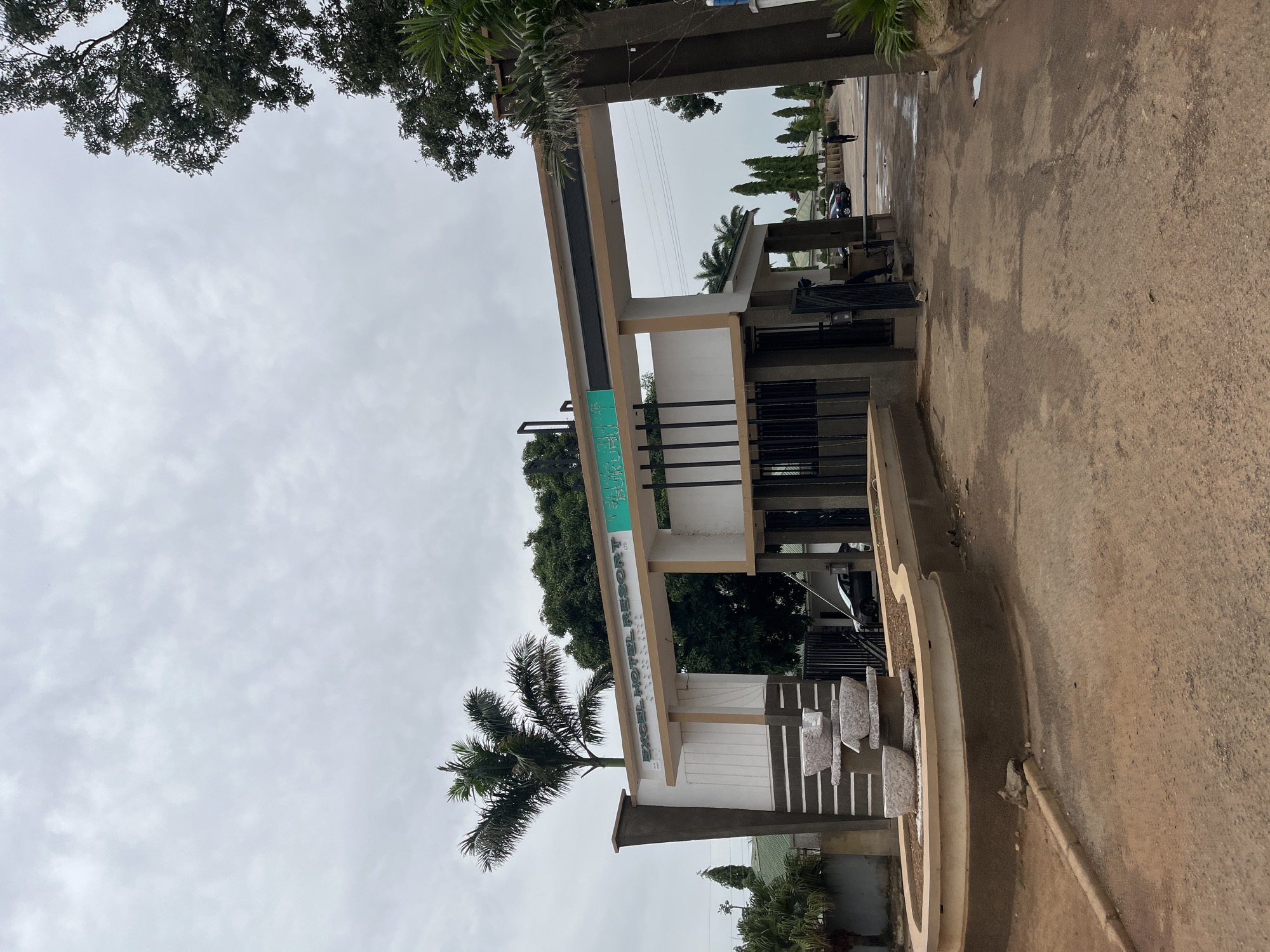 The image shows the entrance to Excel Hotel Resort in Bukuru, Nigeria, under a cloudy sky. The entrance gate is a modern structure with a beige and brown archway. A sign above the archway reads "EXCEL HOTEL RESORT" in white lettering on a beige background, with a green sign to the right that is partially obscured but appears to say "BUKURU".
To the left of the entrance, there is a tiered water fountain made of stone. The ground in front of the entrance is a dirt or gravel road, with some puddles indicating recent rain. Beyond the entrance, several cars are parked, and a few people are walking. There are trees and some buildings visible in the background. The overall scene suggests an outdoor, potentially tropical environment.