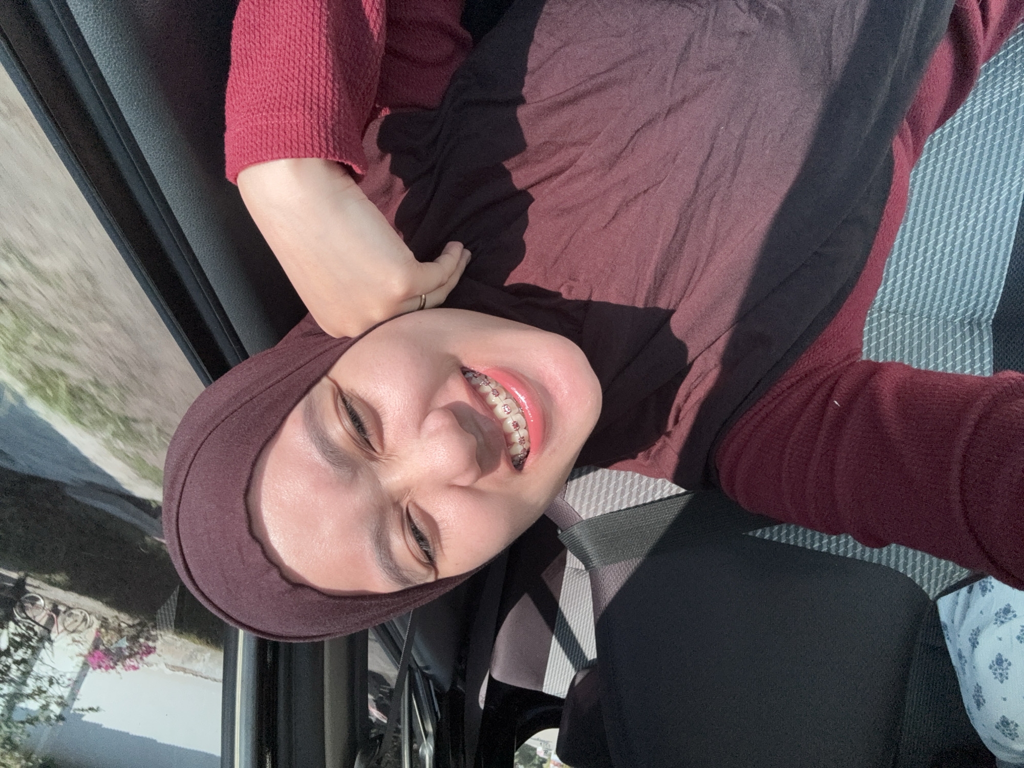 A young woman wearing a maroon hijab and a maroon sweater is taking a selfie inside a vehicle. Her eyes are closed and she is smiling broadly, showing off her braces. Her right hand is resting on her cheek. The interior of the vehicle appears to be black and gray. Through the window, a blurry outdoor scene is visible, suggesting the vehicle is in motion. The background outside the window shows a house with pink flowers and a bicycle. The image appears to be taken during the day. The location is the city of Kebonsari, Indonesia.