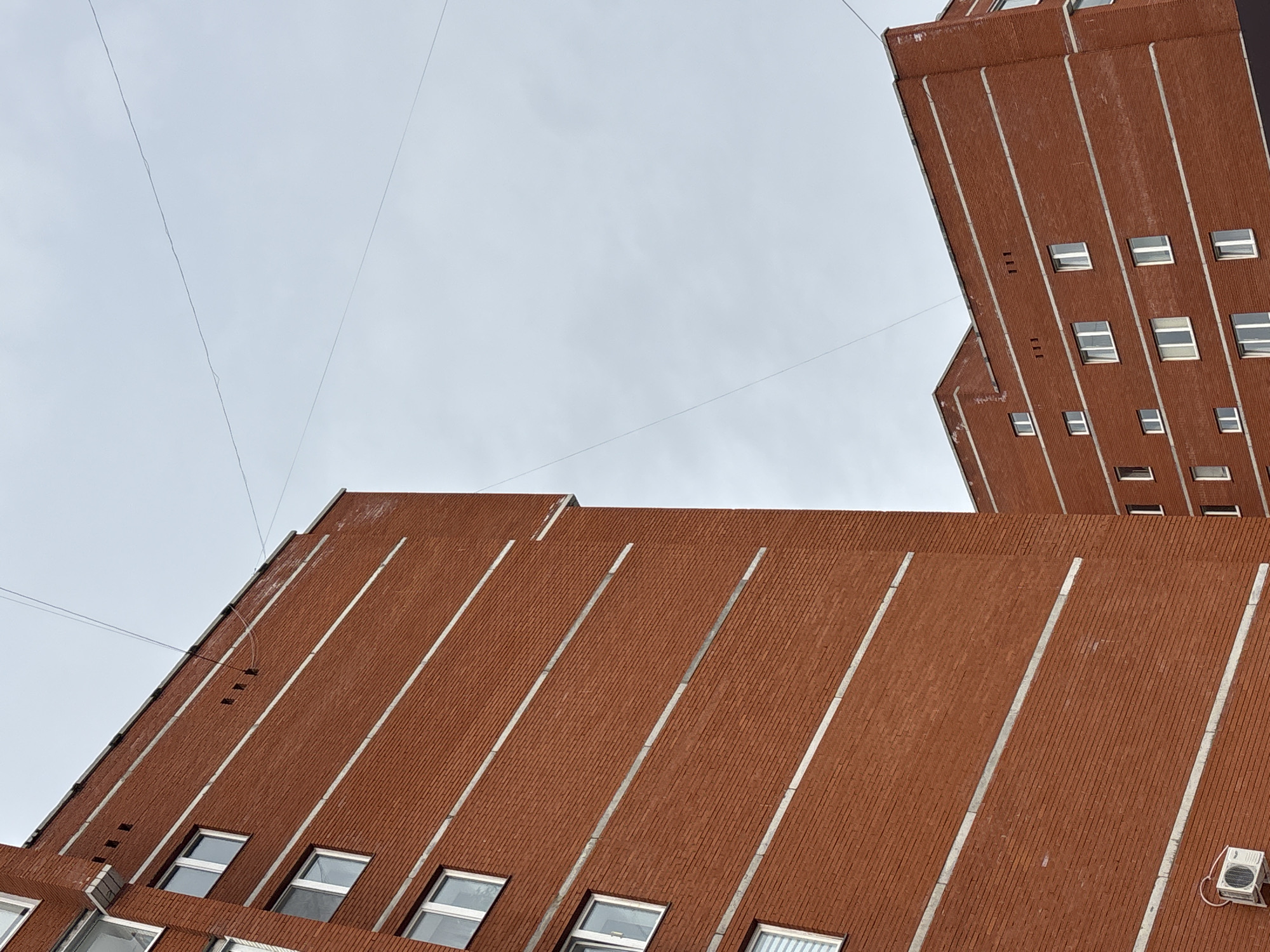 The image displays an urban scene in the city of Chaikovsky, Russia, featuring two multi-story residential or commercial buildings constructed primarily of red brick.
The dominant structure, positioned on the left, is a red brick building with multiple rectangular white-framed windows arranged vertically. Horizontal light grey bands are integrated into the brickwork, delineating floor levels. An external air conditioning unit is mounted on the lower portion of this building's facade. The building has a flat roofline with a parapet, below which several small, dark rectangular openings (possibly vents) are visible. A light grey downspout is present on the upper left edge of the building.
A second, similar red brick building is situated in the mid-ground on the right side of the frame. It also exhibits red brick construction with horizontal light grey bands and numerous white-framed rectangular windows. Its flat roofline and parapet are also discernible.
The sky is uniformly overcast and light grey. Multiple thin, dark utility wires traverse the sky horizontally and diagonally, connecting across the scene between the structures. No individuals or specific actions are visible.
